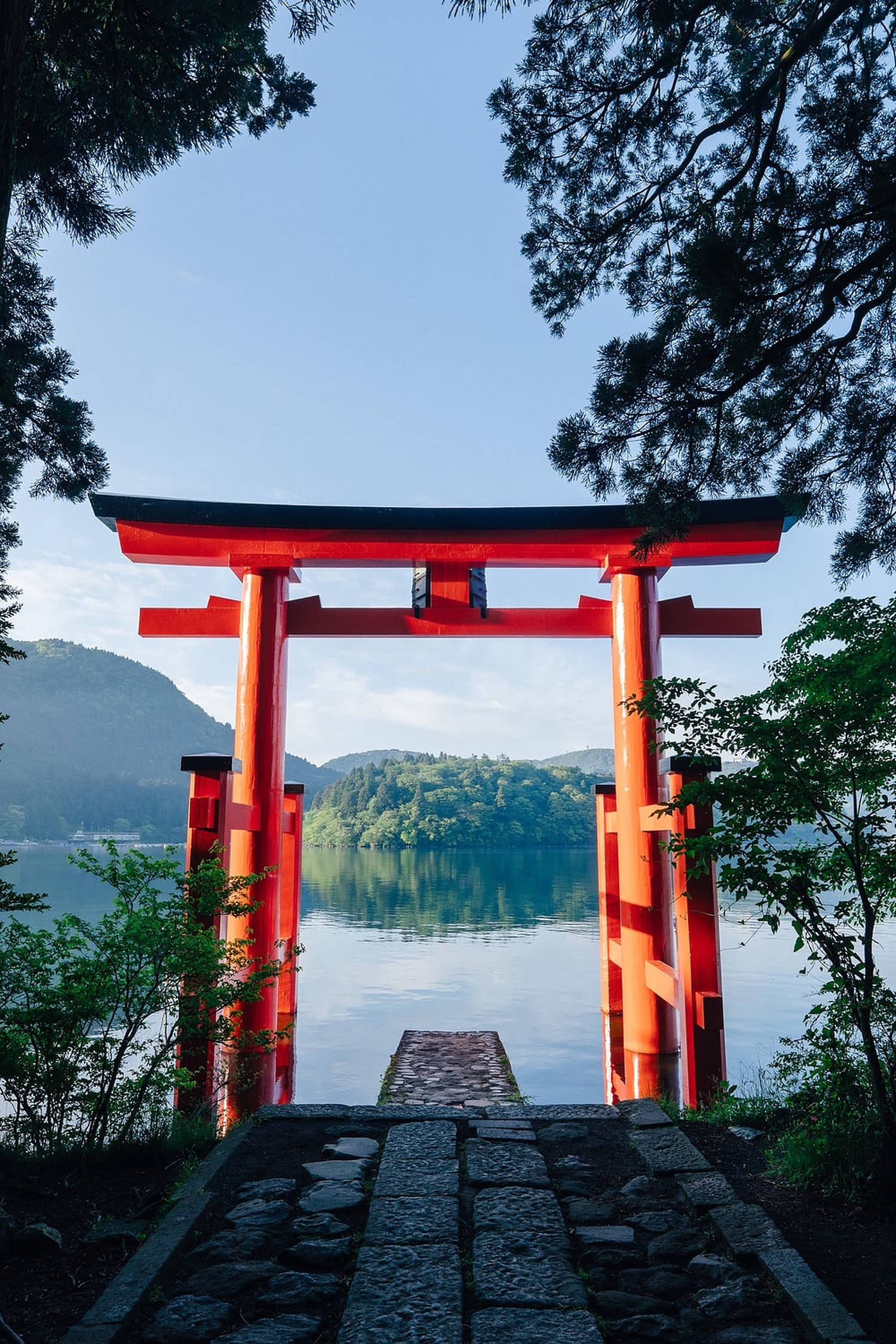 Torii 桌布 壁紙 — 一座鮮紅色的鳥居矗立在寧靜的湖邊，四周綠樹成蔭，天空湛藍。