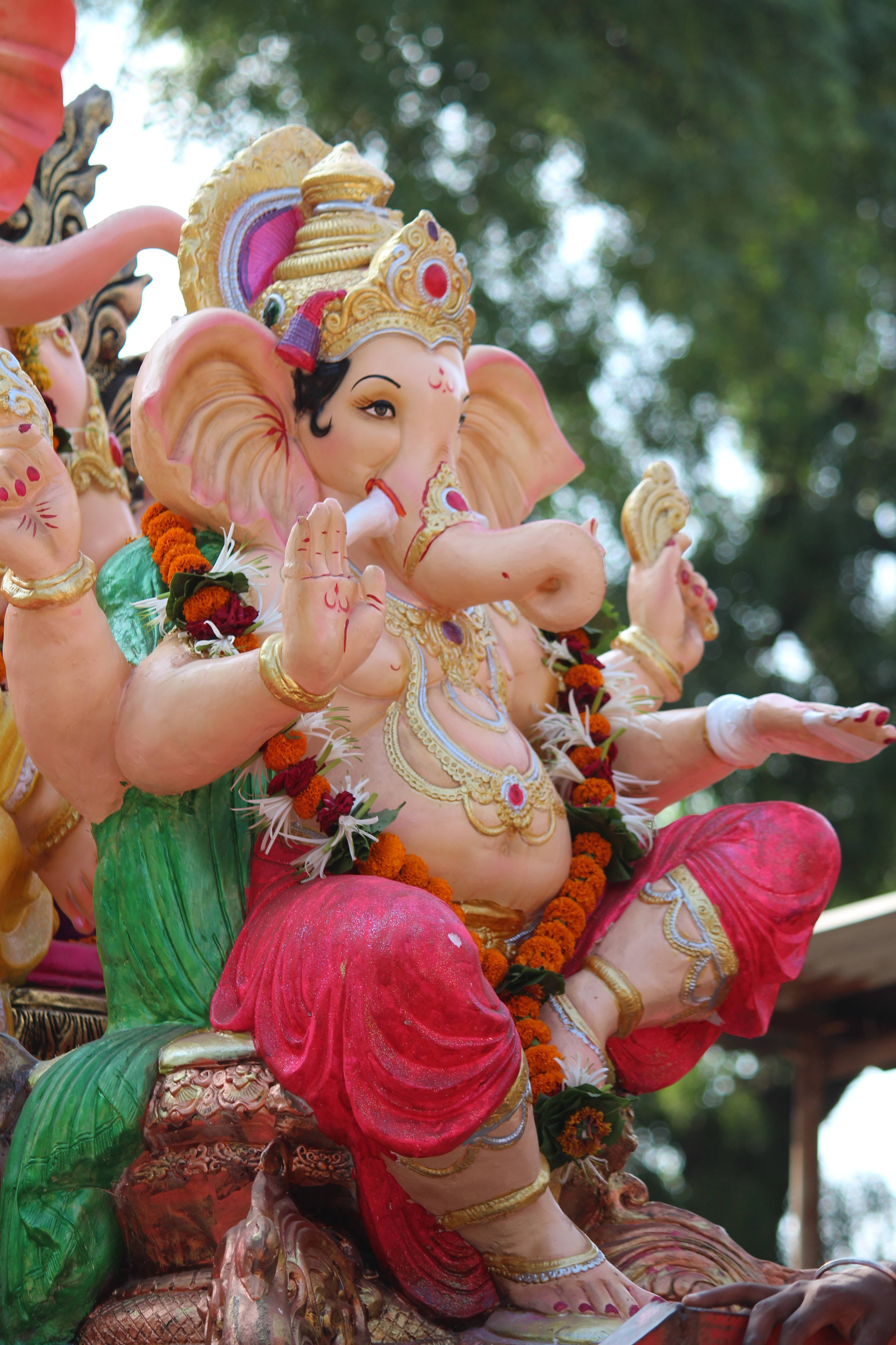 ganesh 4K 桌布 壁紙 — 一座鮮豔的粉紅色象頭神雕像，飾有黃金首飾和萬壽菊花環，靜靜地坐在綠色樹葉中。