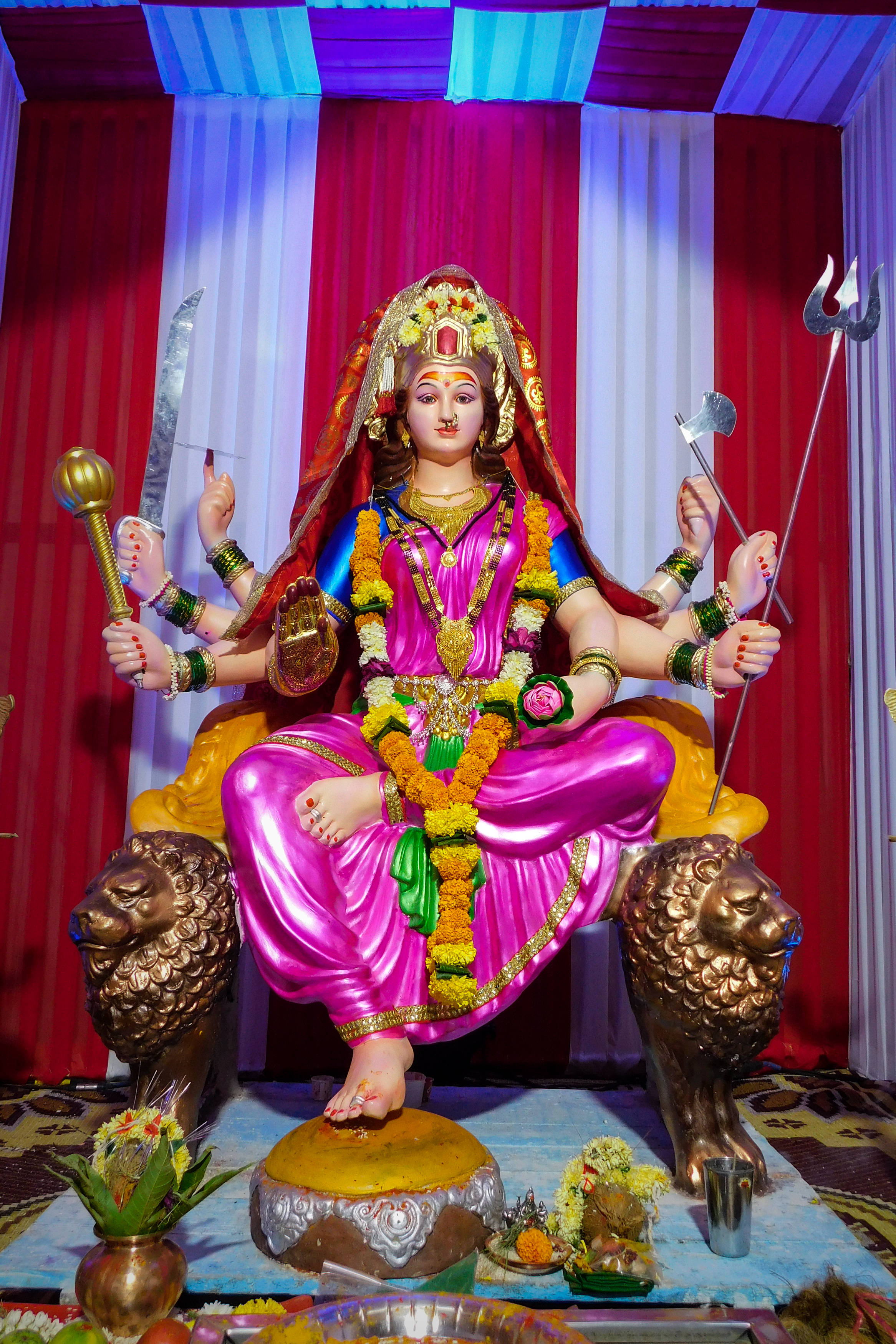 Navratri 桌布 壁紙 — 一座鮮豔粉紅色、多臂的女神雕像，飾有黃金珠寶和花環，莊嚴地坐在獅子寶座上，背景是紅色和白色的窗簾。