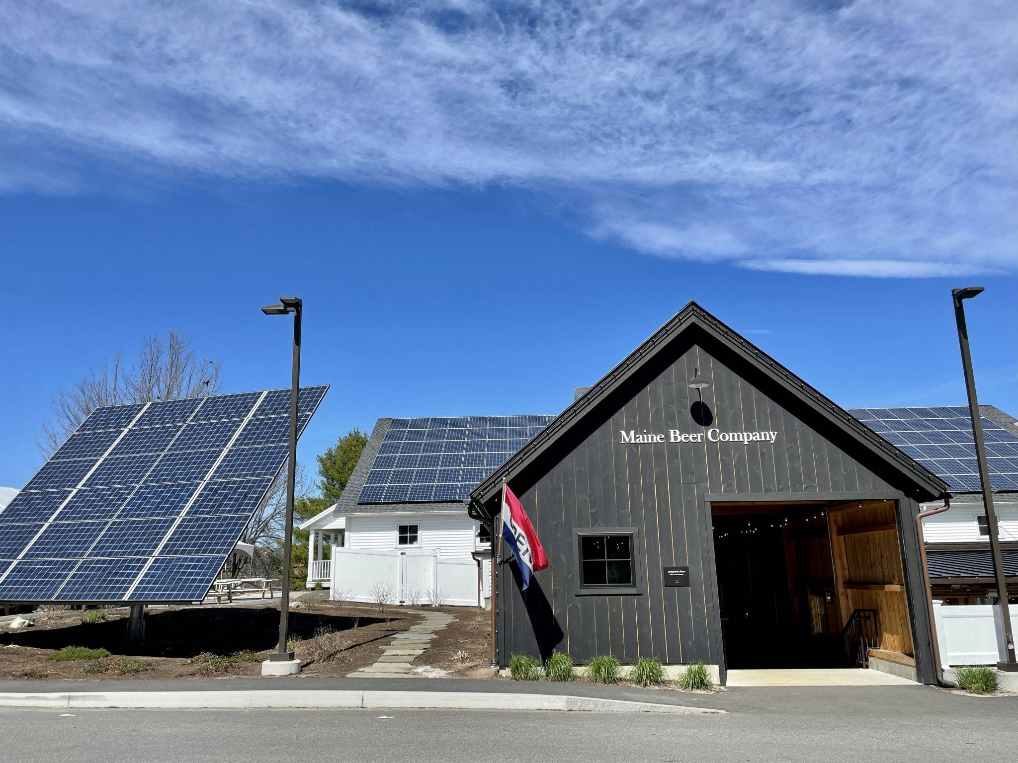 Building 桌布 壁紙 — 一座黑色木造建築物，標有 Maine Beer Company，屋頂上裝有太陽能板，附近還有一個大型傾斜面板，在明亮的藍天下。