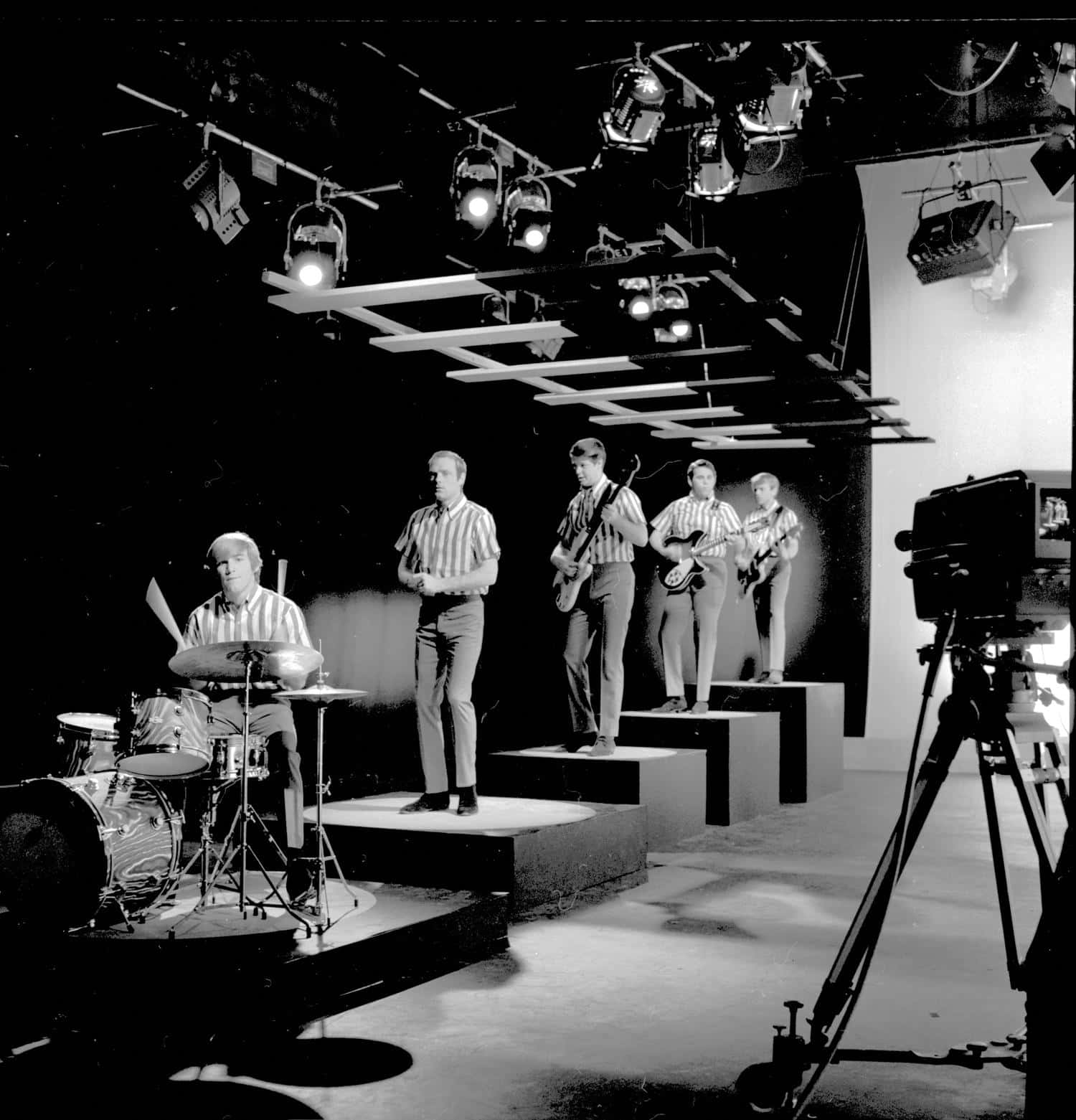 band 桌布 壁紙 — 一張1960年代樂團在分層舞台上、在攝影棚燈光下、相機放在三腳架上表演的黑白照片。
