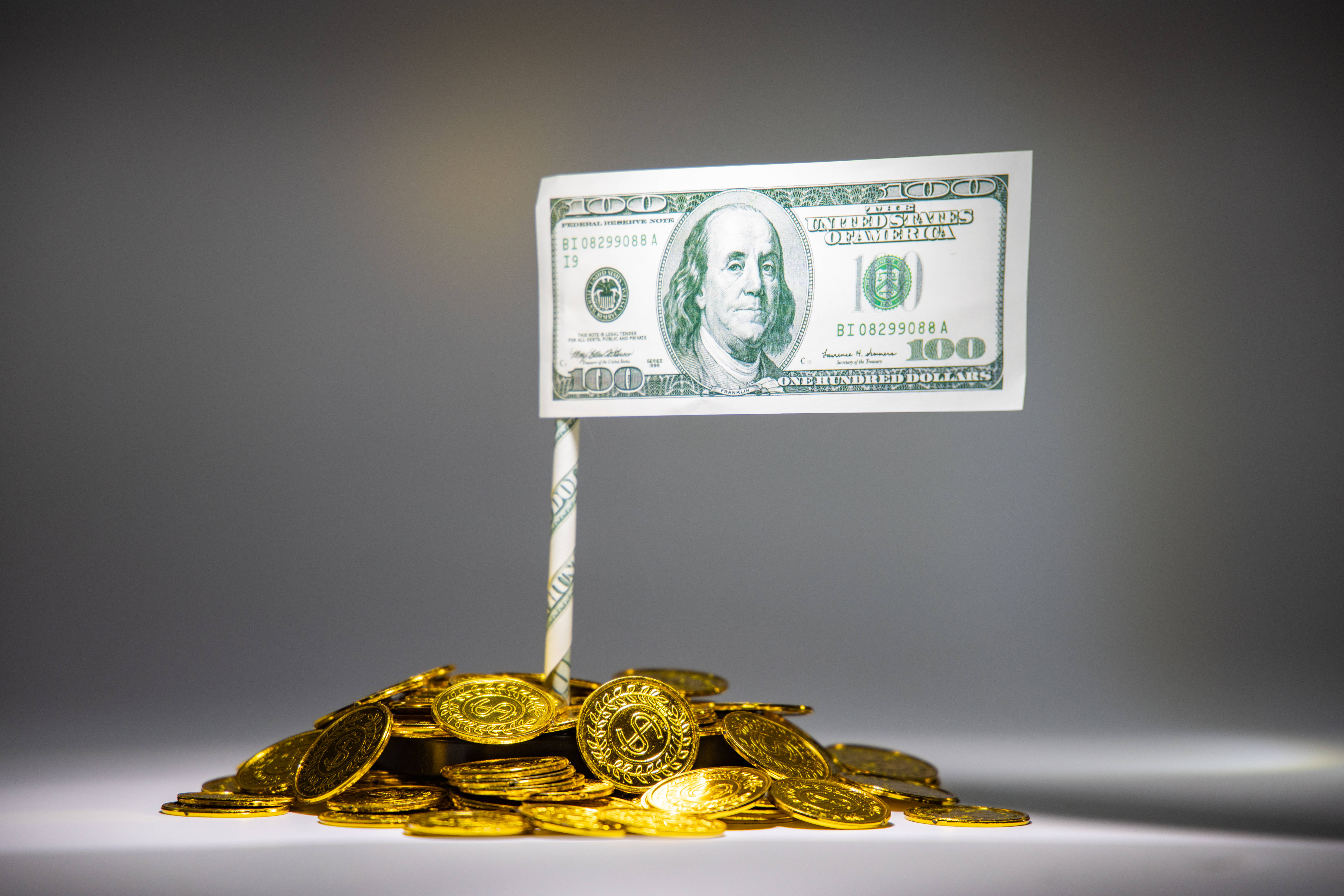 金錢 桌布 壁紙 — 一張一百美元紙幣豎立在一堆金幣中，背景為漸變灰色。