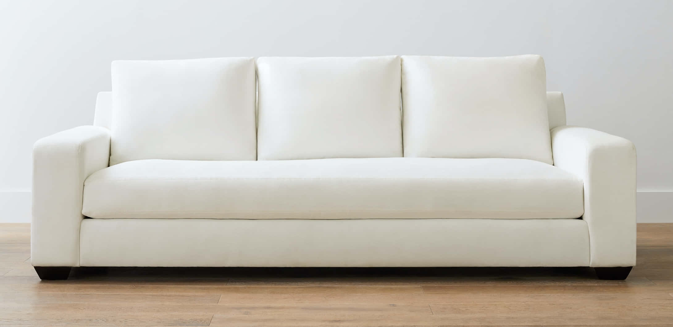 Couch 桌布 壁紙 — 一張乾淨簡約的白色現代沙發，配有三個靠墊，置於淺色木地板的中央，背景是純白牆壁。