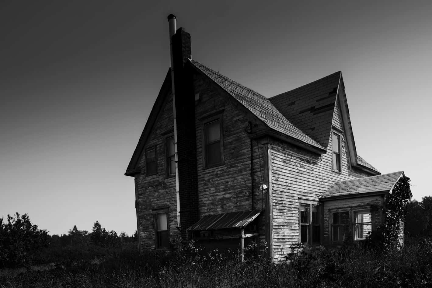 Haunted House 桌布 壁紙 — 一張令人毛骨悚然的黑白照片，拍攝了一座風化的木屋，孤零零地矗立在荒蕪的田野中，天空陰沉。