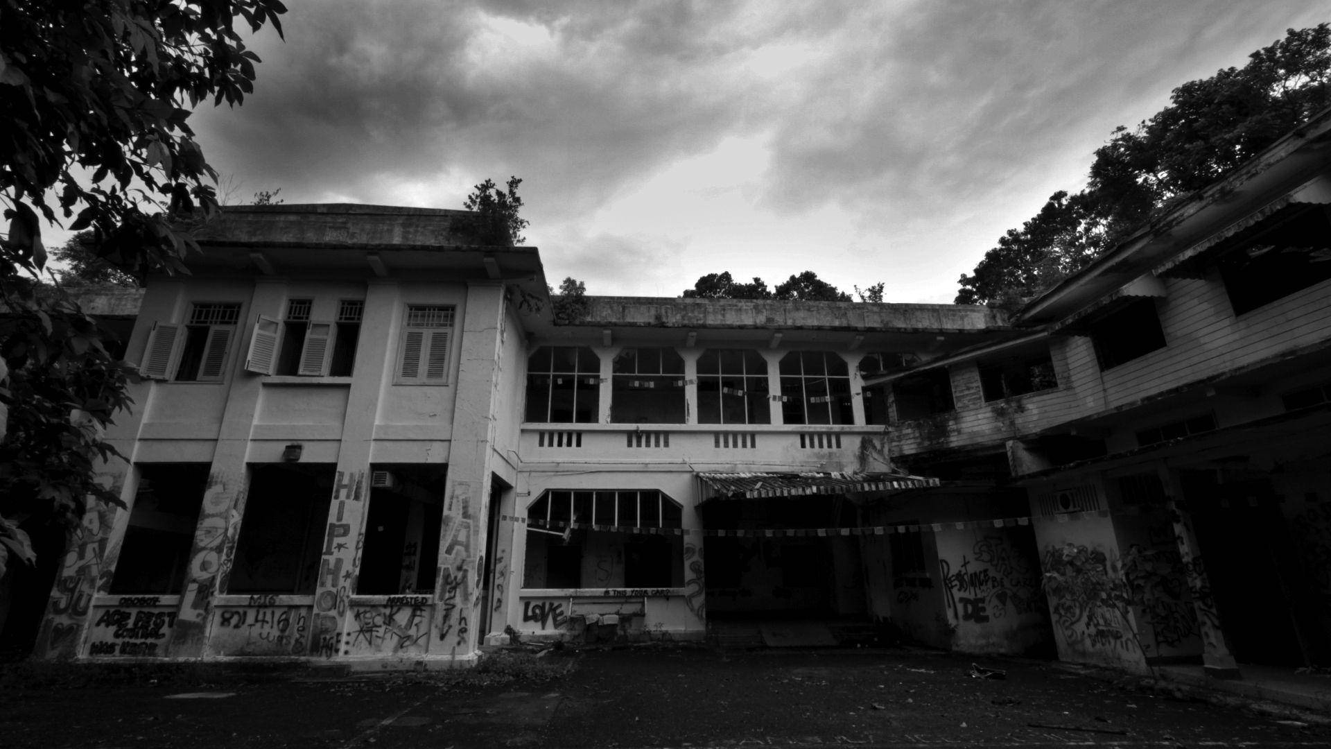 Haunted Mansion 桌布 壁紙 — 一張令人毛骨悚然的黑白照片，拍攝了一座破敗的殖民風格建築，藤蔓叢生，塗鴉遍佈，背景是風暴天空。