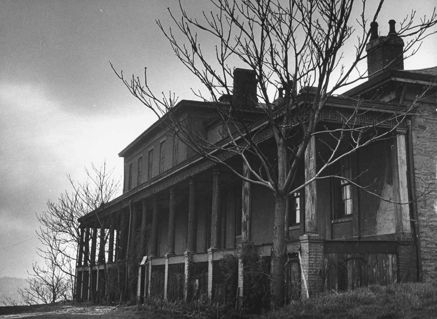 Haunted House 桌布 壁紙 — 一張令人毛骨悚然的黑白照片，拍攝了一棟破舊的兩層房屋，光禿禿的樹木框住了風化的門廊。