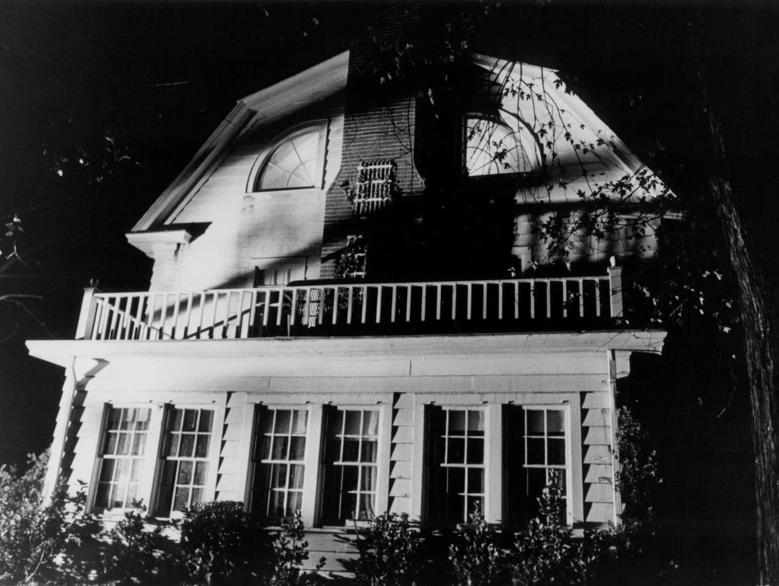 Haunted House 桌布 壁紙 — 一張令人難忘的黑白夜景照片，拍攝了一棟帶有門廊和拱形窗戶的兩層房屋，部分被樹木遮擋。