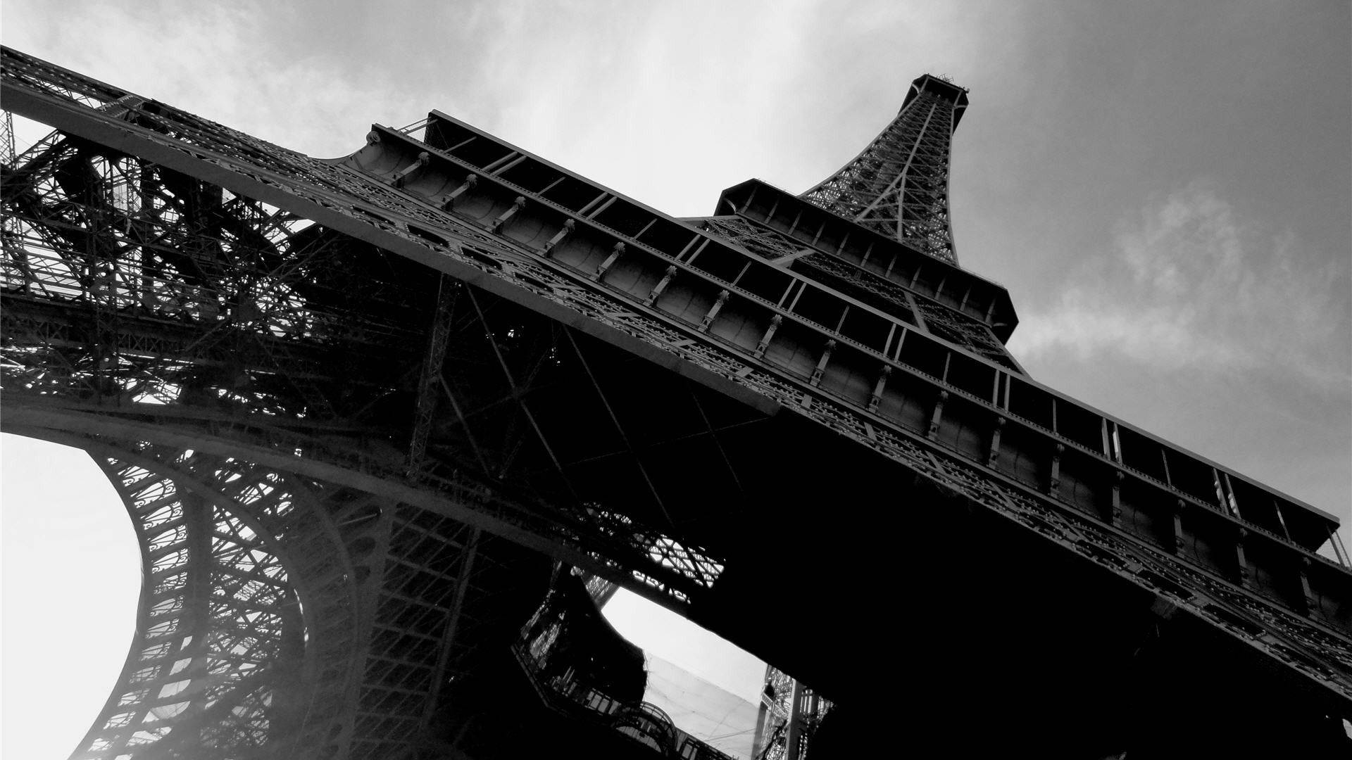Eiffel Tower 桌布 壁紙 — 一張以低角度拍攝的戲劇性黑白照片，展現艾菲爾鐵塔複雜的鐵格子結構，背景是陰雲密布的天空。