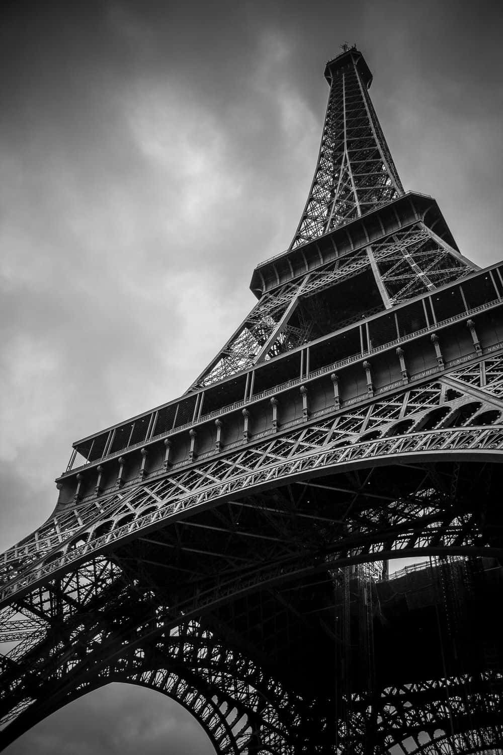 Black And White Architecture 桌布 壁紙 — 一張以低角度拍攝的戲劇性黑白照片，呈現艾菲爾鐵塔複雜的鐵製格架，背景是陰雲密佈的天空。
