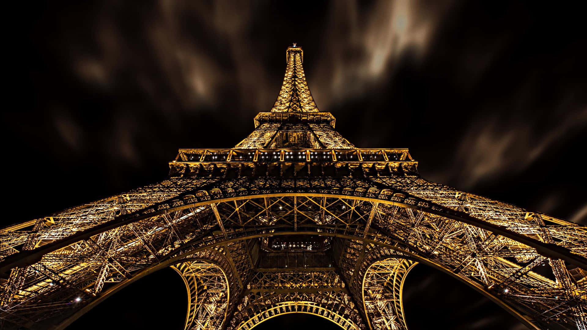 Eiffel Tower 桌布 壁紙 — 一張低角度拍攝的戲劇性夜景，艾菲爾鐵塔在黑暗多雲的天空中閃耀著金色光芒。