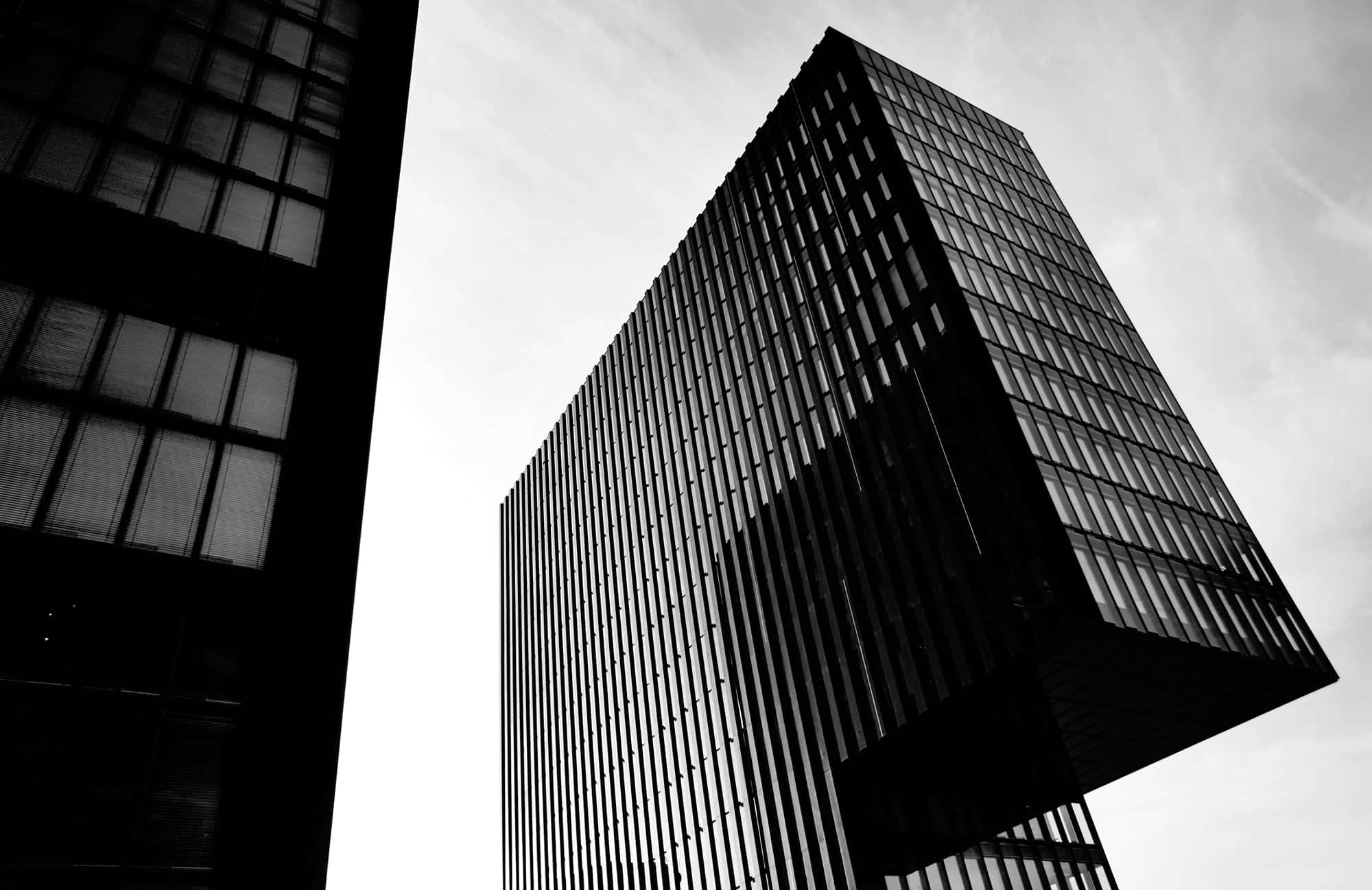 Black And White City 桌布 壁紙 — 一張低角度拍攝的戲劇性黑白照片，展現兩座具有銳利幾何線條的現代摩天大樓，背景是多雲的天空。