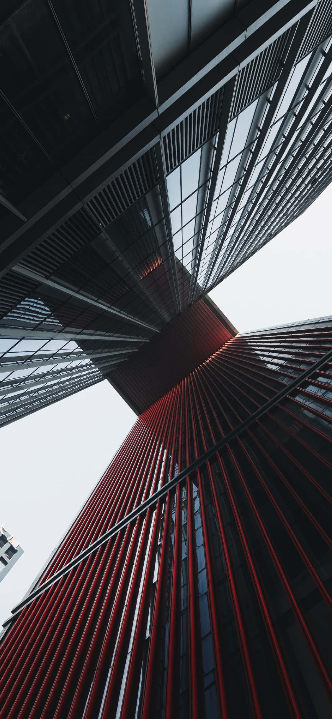 建築 桌布 壁紙 — 一張低角度拍攝的現代摩天大樓戲劇性照片，建築物帶有醒目的紅色垂直條紋和玻璃面板，伸向蒼白的天空。