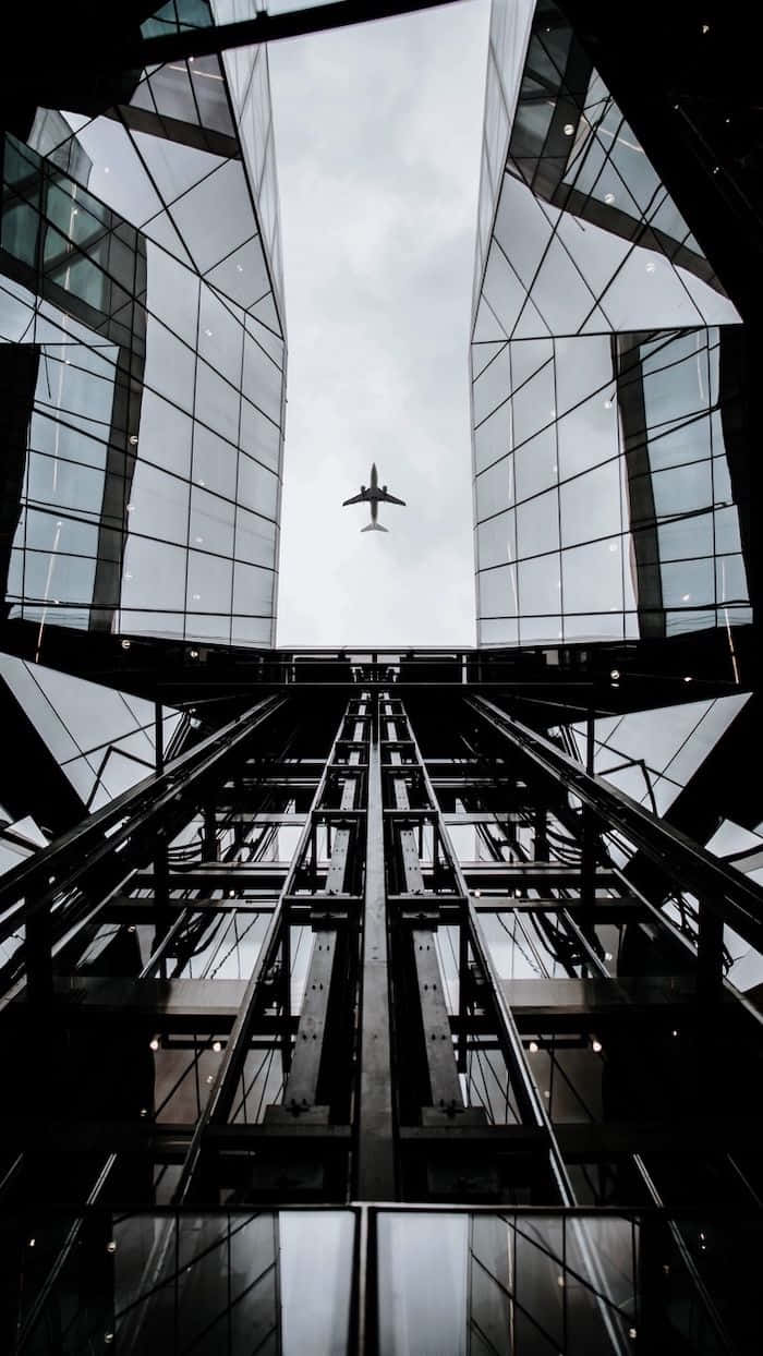 Plane 桌布 壁紙 — 一張低角度黑白照片，拍攝飛機在擁有反光外牆的 towering 玻璃摩天大樓之間飛行。