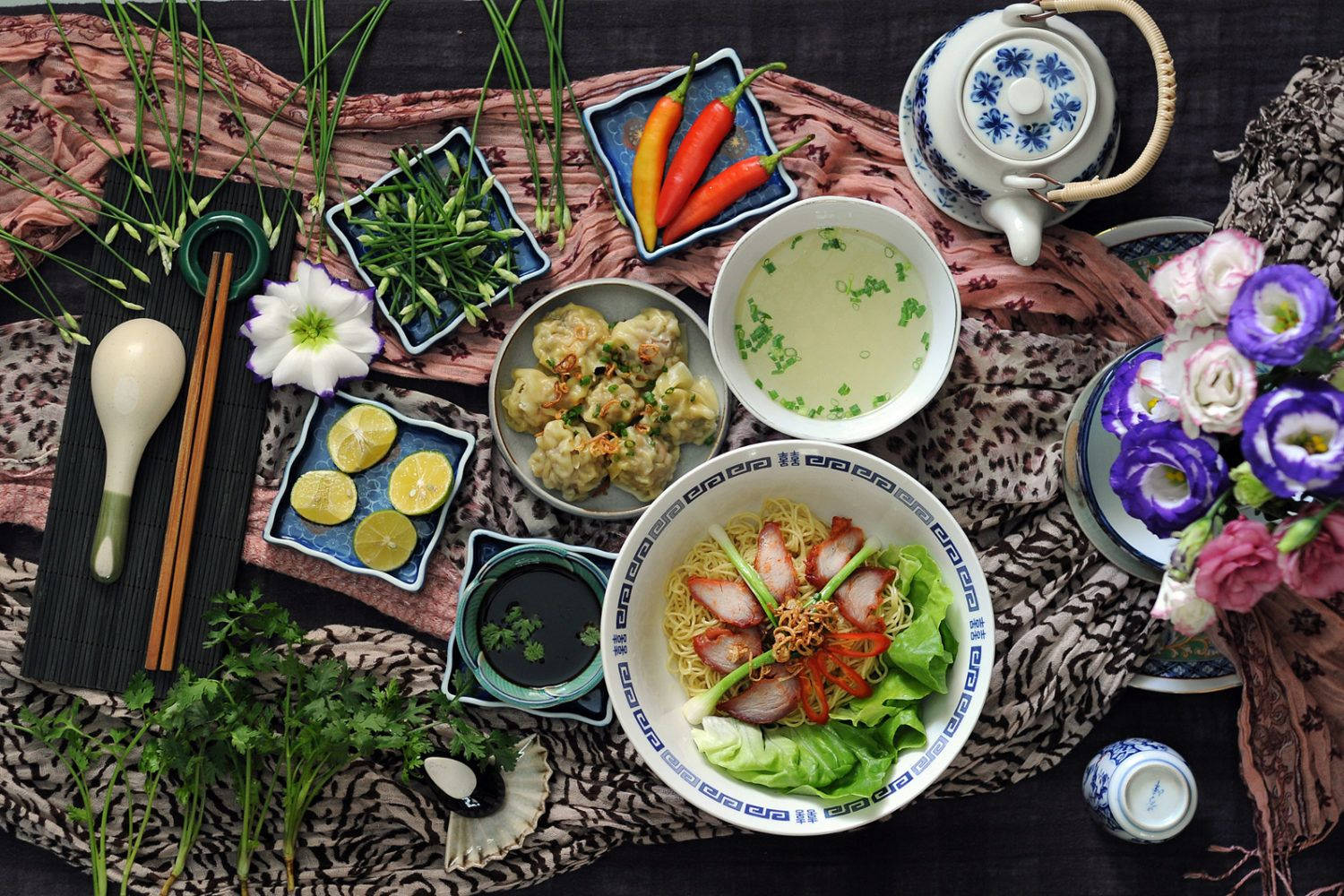bowl 桌布 壁紙 — 一張充滿活力的亞洲美食俯拍照片，有麵條、餃子、湯、辣椒和花朵，擺放在圖案布上。