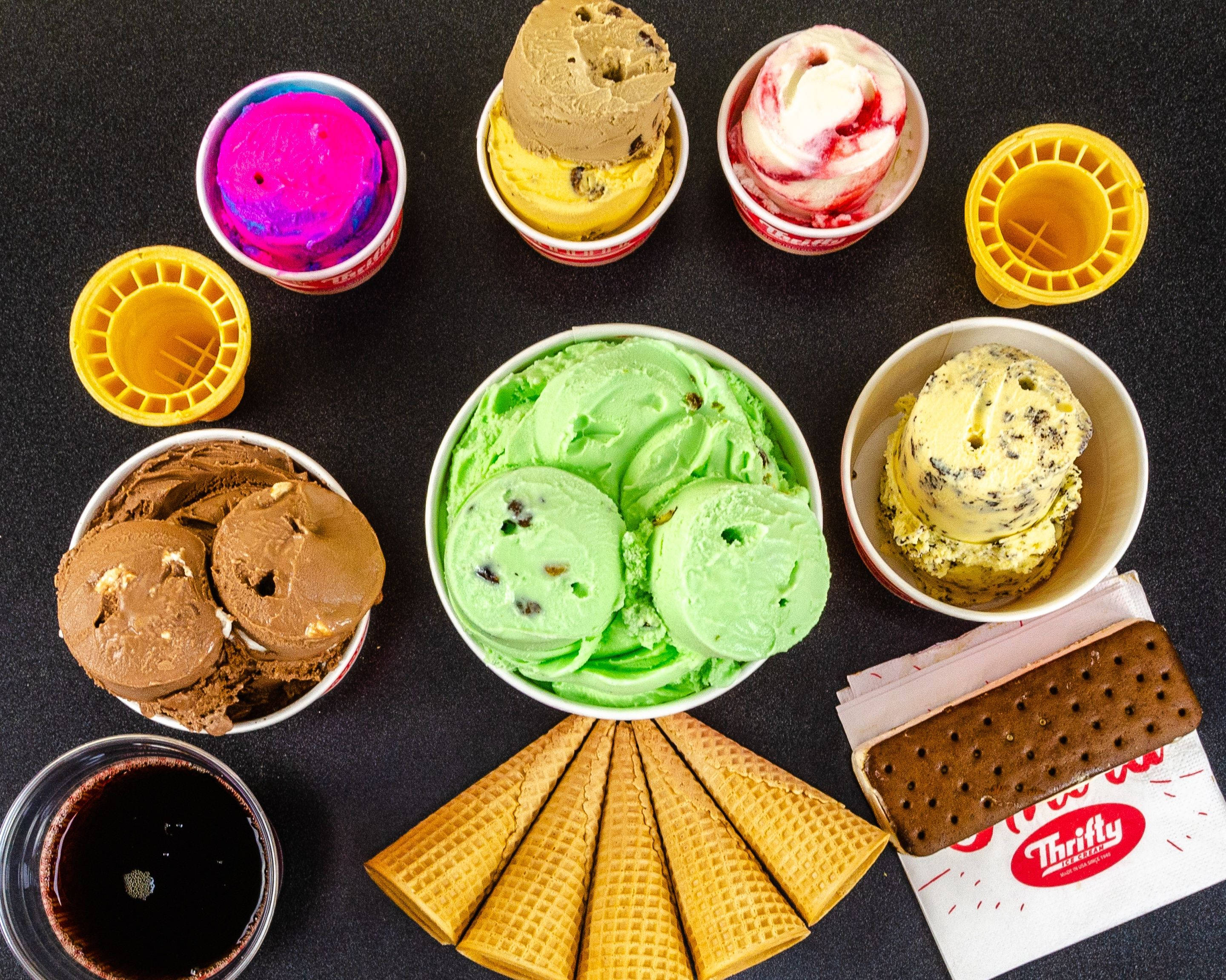 Ice Cream 桌布 壁紙 — 一張充滿活力的俯拍照片，展示了各種色彩繽紛的冰淇淋球、碗、甜筒和一杯巧克力飲品，排列在深色表面上。