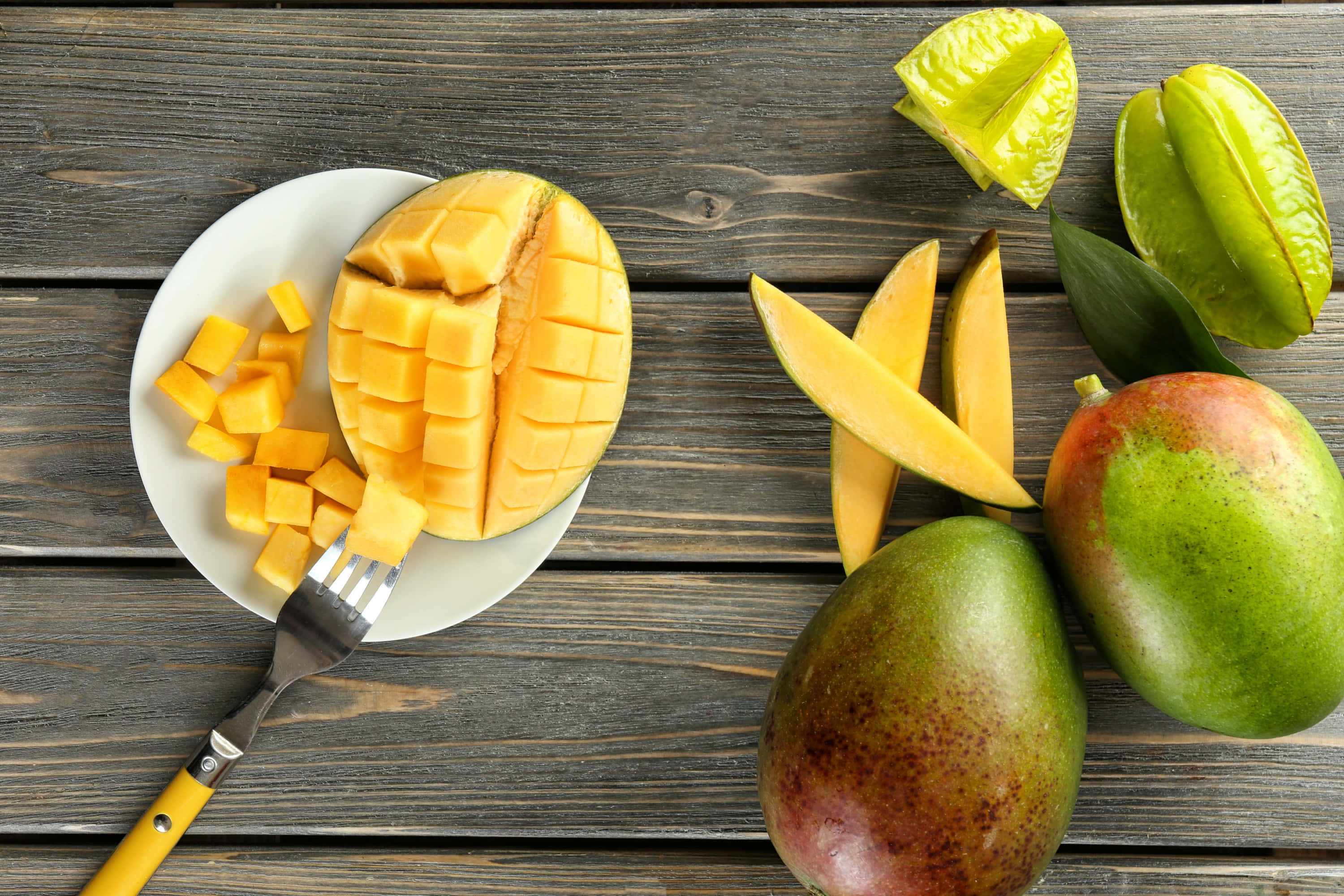 Mango 桌布 壁紙 — 一張充滿活力的俯拍照片，展示切片芒果和楊桃放在質樸的木桌上，旁邊有一把叉子。