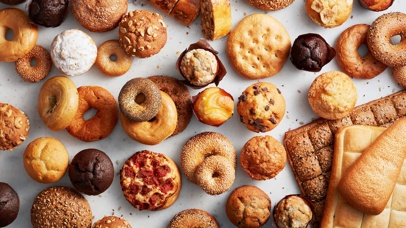 1366x768 Pastries 桌布 壁紙 — 一張充滿活力的俯拍照片，展示各種烘焙食品，包括貝果、馬芬和麵包，散落在白色表面上。