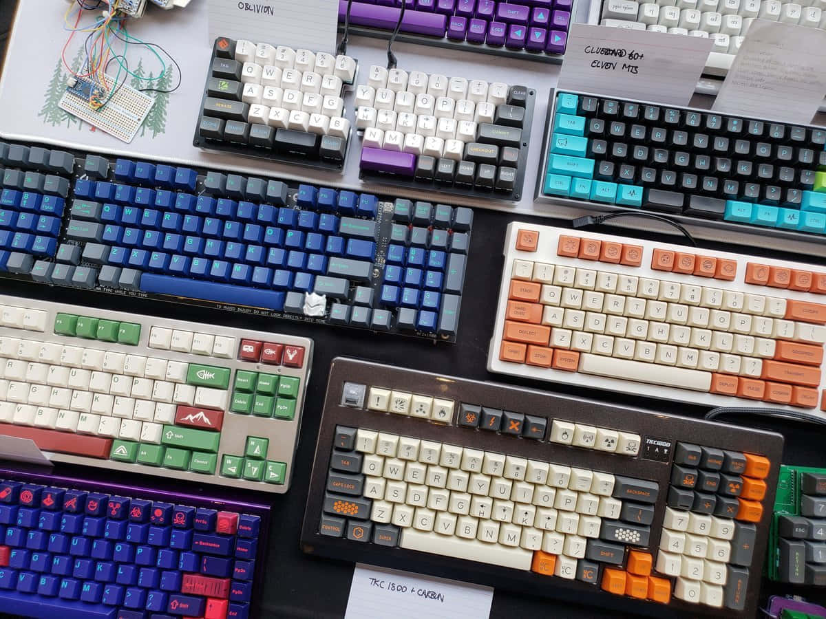 Cute Keyboard 桌布 壁紙 — 一張充滿活力的俯拍照片，展示各種帶有彩色鍵帽和自訂佈局的機械鍵盤，排列在深色表面上。