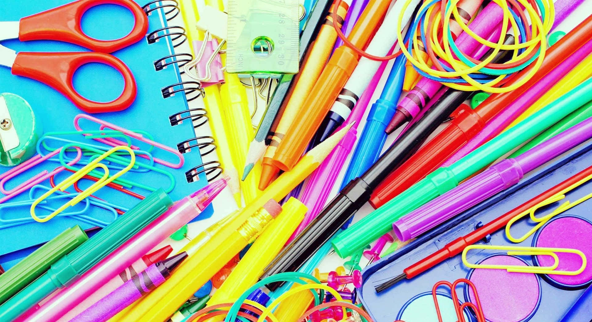 Cool School 桌布 壁紙 — 一張充滿活力的俯拍照片，展示散落在藍色筆記本上的各種彩色文具，包括筆、剪刀和迴紋針。