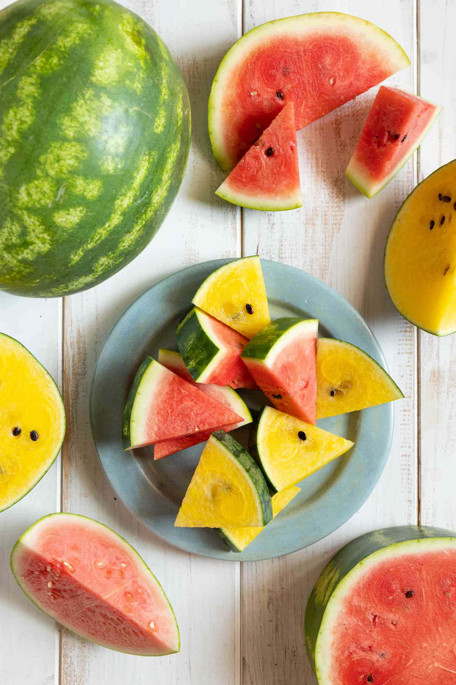 Watermelon 桌布 壁紙 — 一張充滿活力的俯拍照片，展示紅色和黃色西瓜片排列在盤子上，並散落在白色木質表面上的整個西瓜周圍。