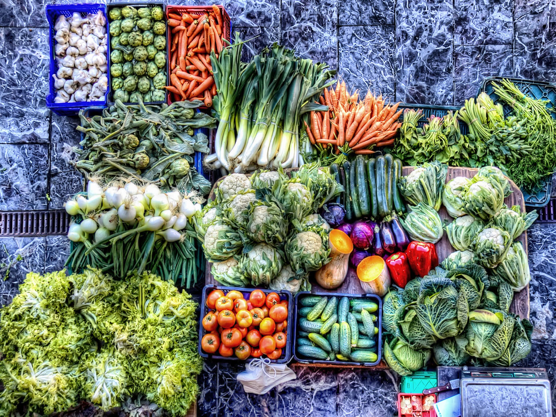 Vegetables 桌布 壁紙 — 一張充滿活力的俯拍照片，展示胡蘿蔔、番茄和花椰菜等新鮮蔬菜，整齊排列在深色磁磚市場攤位的彩色板條箱中。