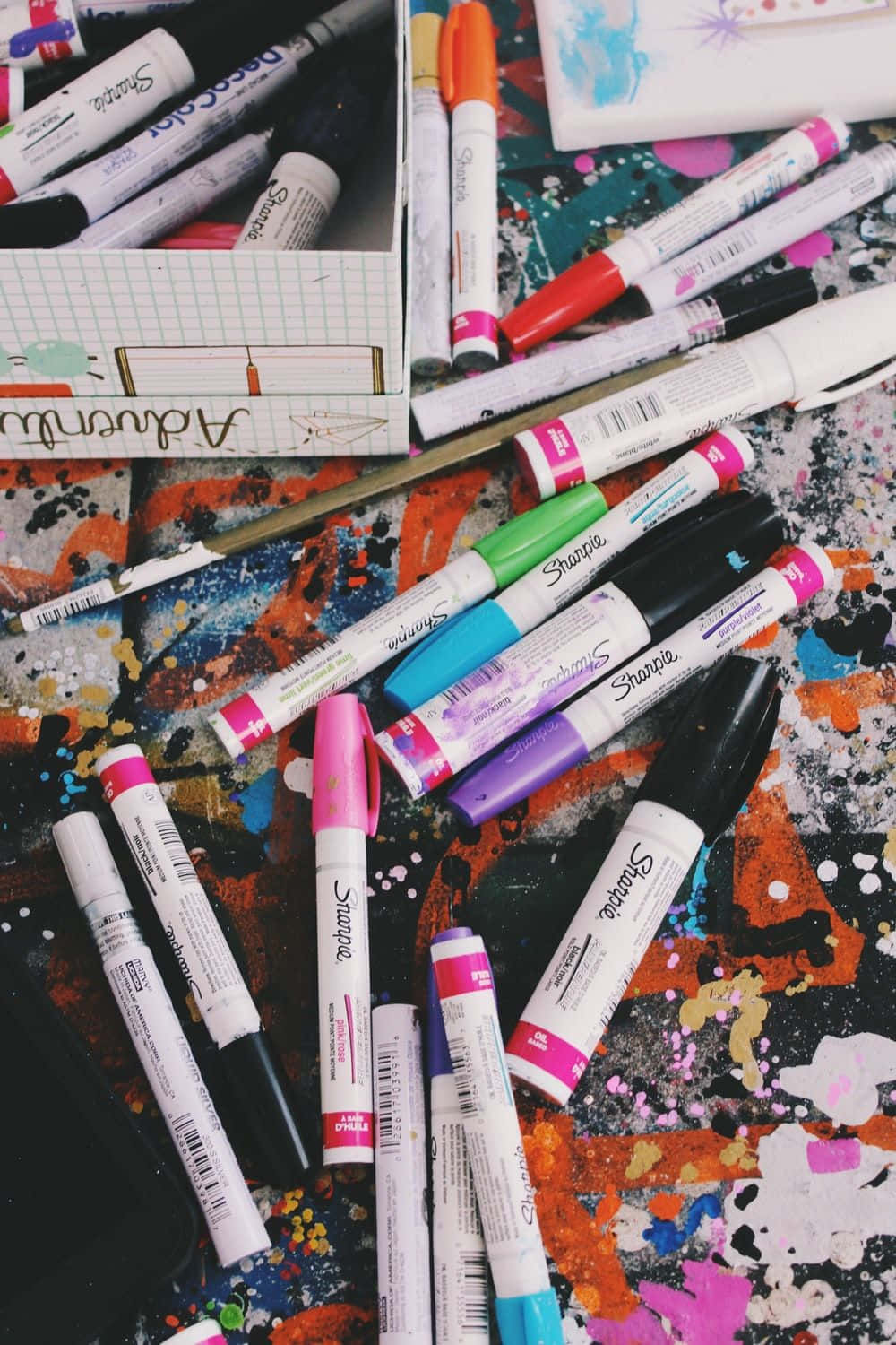Paint Splatter Aesthetic 桌布 壁紙 — 一張充滿活力的俯拍照片，展示色彩繽紛的Sharpie記號筆散落在濺滿油漆的藝術表面上，旁邊還有一本筆記本。