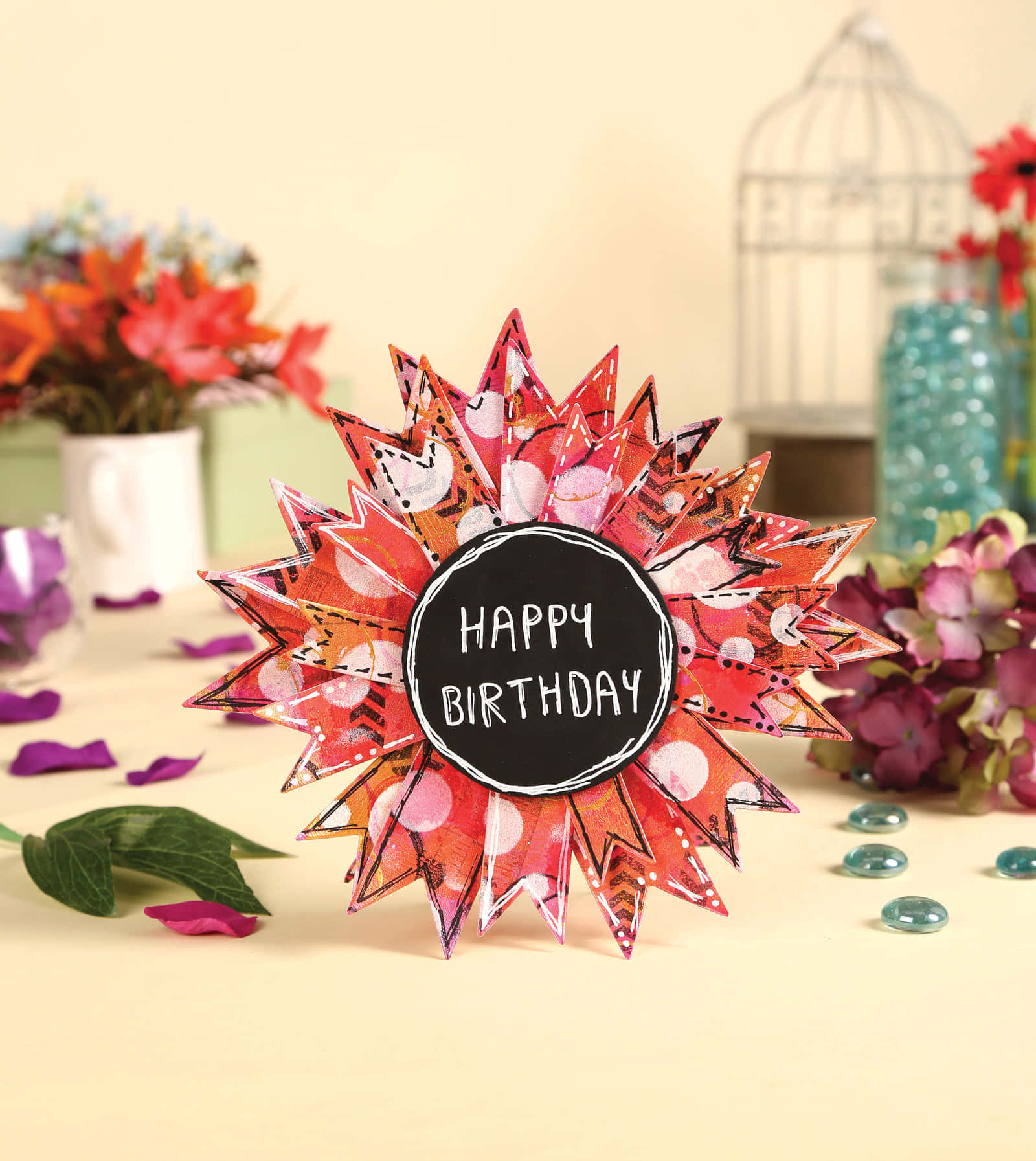Fall Birthday 桌布 壁紙 — 一張充滿活力的手工生日卡，形狀像太陽爆發，帶有紅色圖案紙花瓣和一個黑色黑板中心，上面寫著「HAPPY BIRTHDAY」，放在一張被鮮花和裝飾玻璃珠環繞的桌子上。