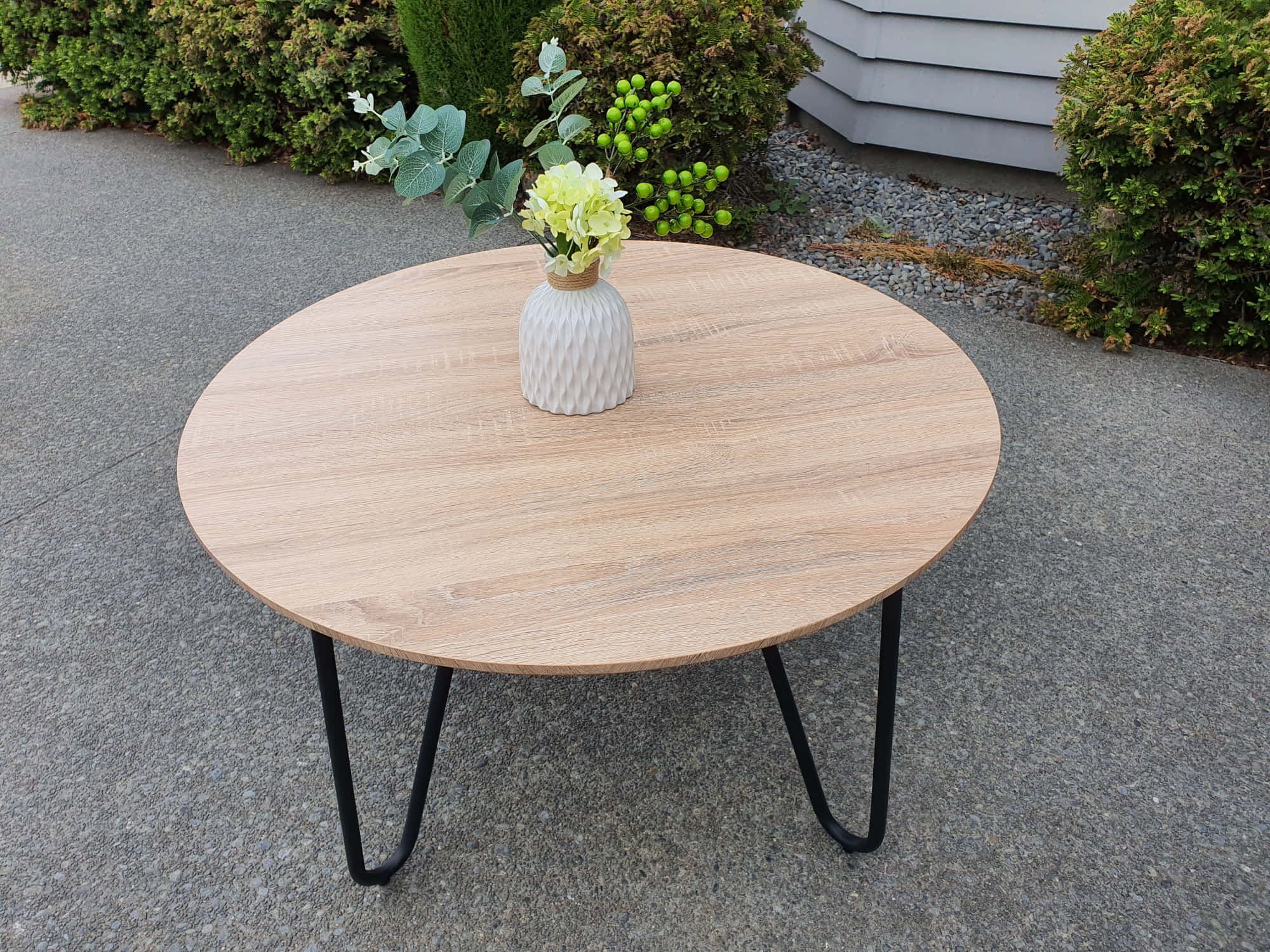 table 桌布 壁紙 — 一張圓形木製咖啡桌，帶黑色髮夾腿，在灰色露臺上放著一個白色花瓶，裡面有綠色植物和鮮花。