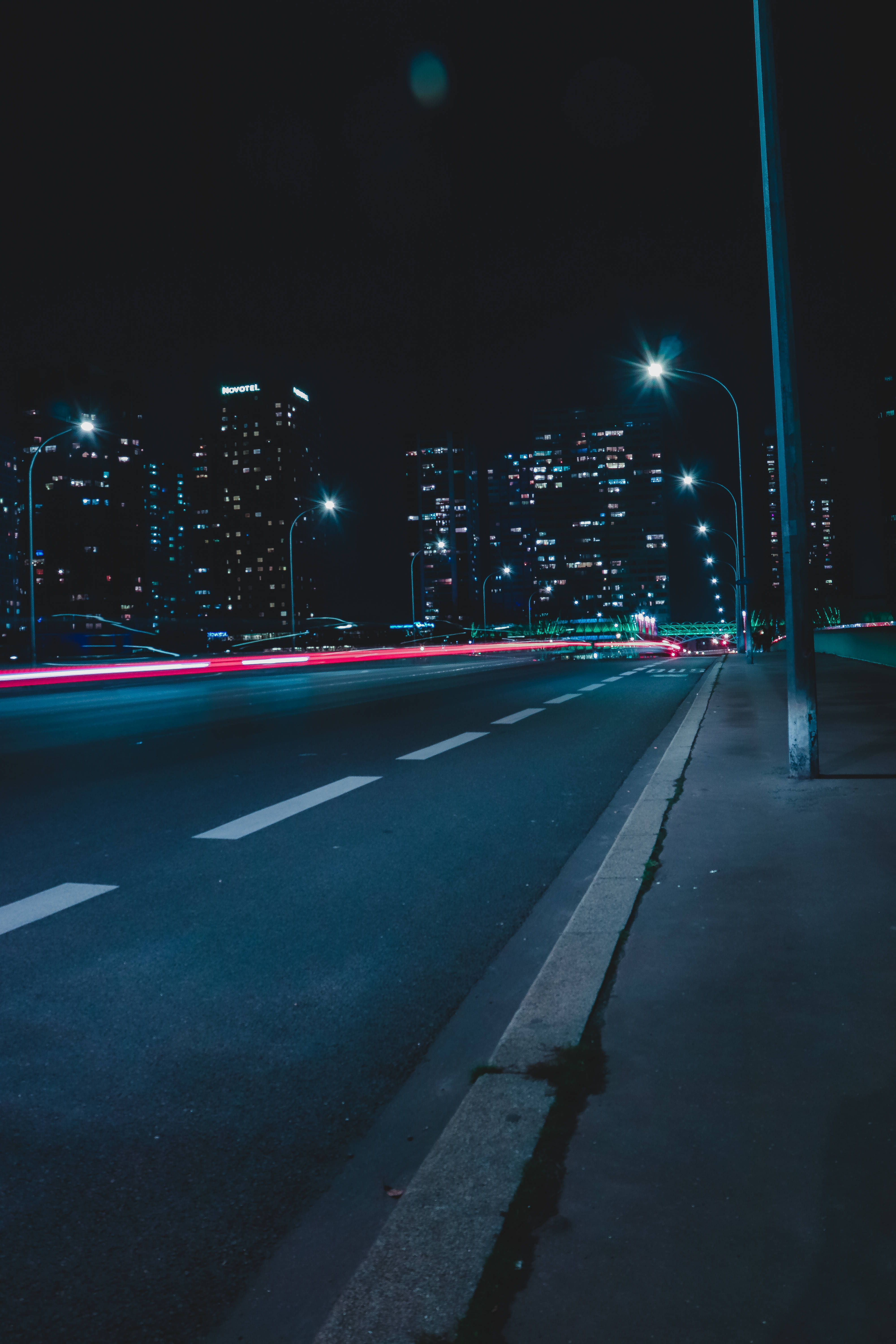 Road 桌布 壁紙 — 一張城市街道的夜間長曝光照片，顯示出紅色尾燈的條紋和發光的街燈，背景是黑暗的摩天大樓。