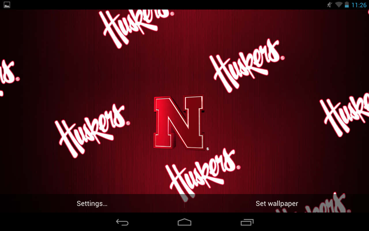 桌布 壁紙 — 一張大膽的紅色壁紙，中央有 Huskers 'N' 標誌，周圍散落著白色草書 Huskers 文字，背景為有紋理的深紅色。