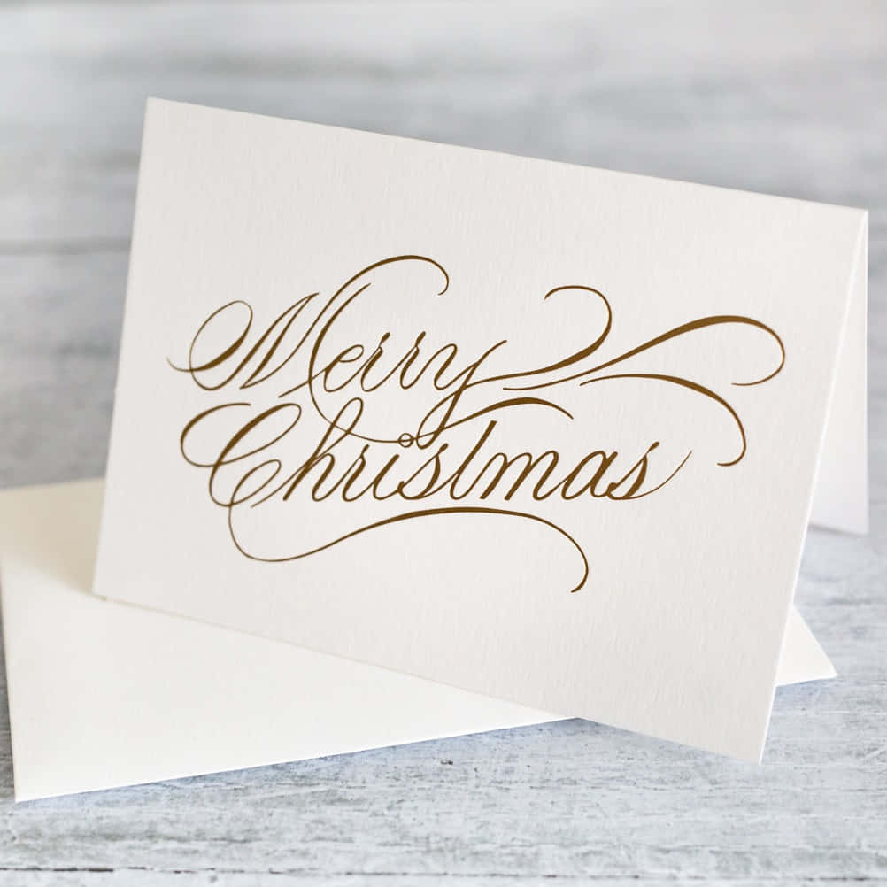 Christmas Card 桌布 壁紙 — 一張奶油色卡片上優雅的金色書法寫著「聖誕快樂」，放在白色信封上，背景是風化的灰色木質表面。