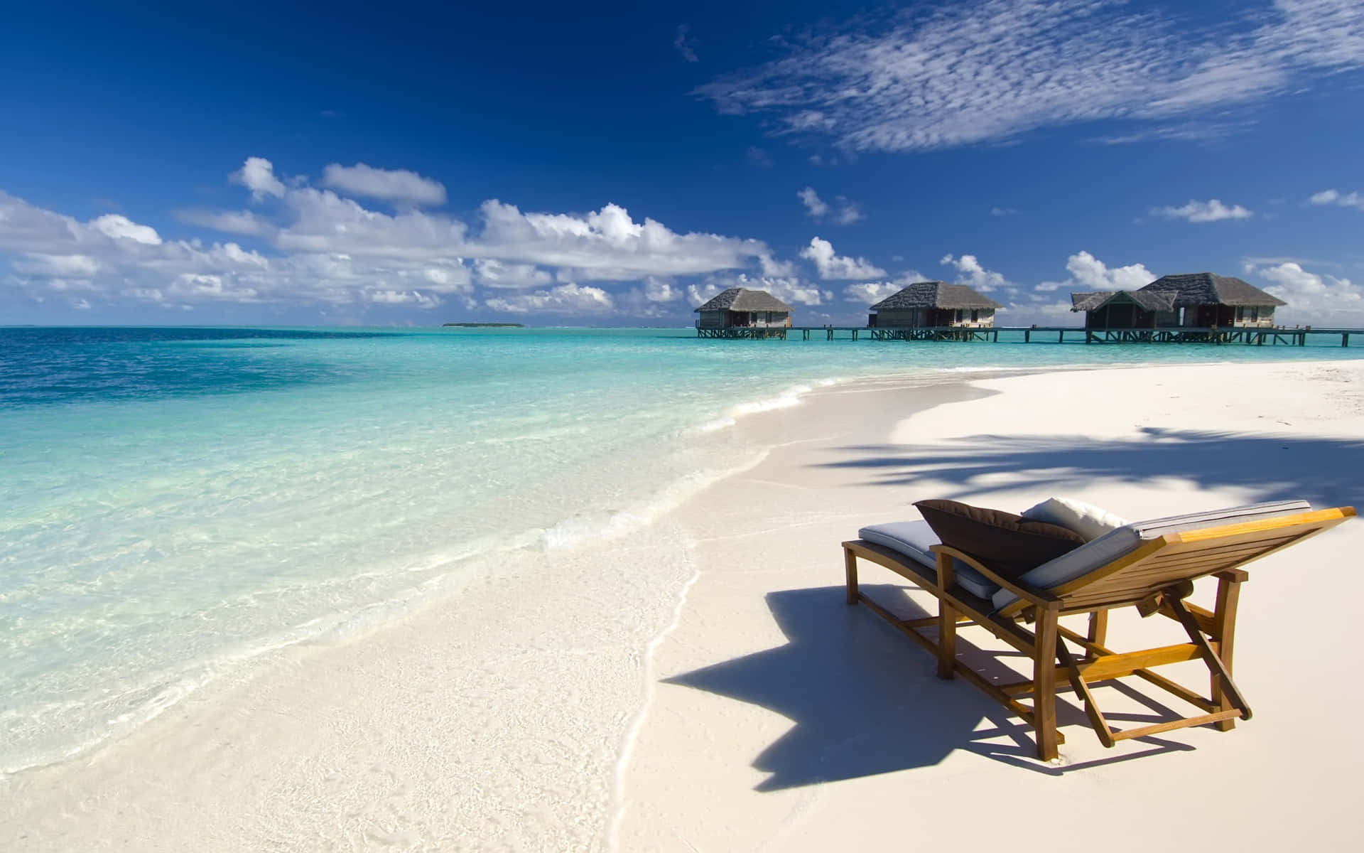 Relaxing Desktop 桌布 壁紙 — 一張孤獨的木製躺椅放在純白沙灘上，面向碧綠的海水，遠處有水上平房，頭頂是明亮的藍天。