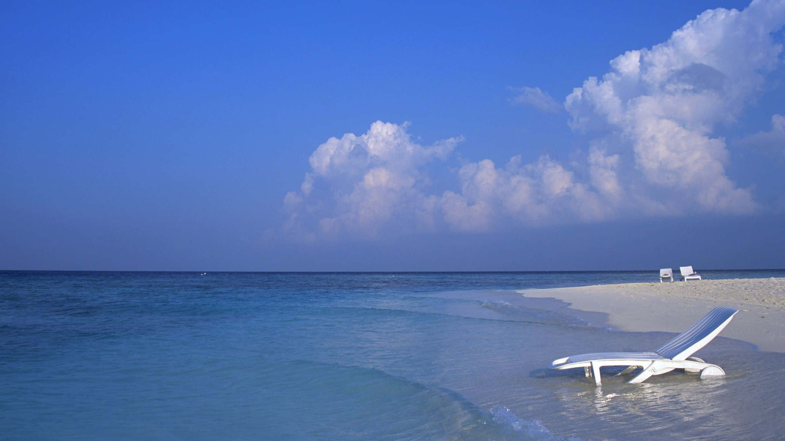 1440p Beach 桌布 壁紙 — 一張孤獨的白色躺椅擱在淺淺的藍綠色海水中，位於純淨的白色沙灘上，上方是廣闊的藍天和蓬鬆的雲朵。
