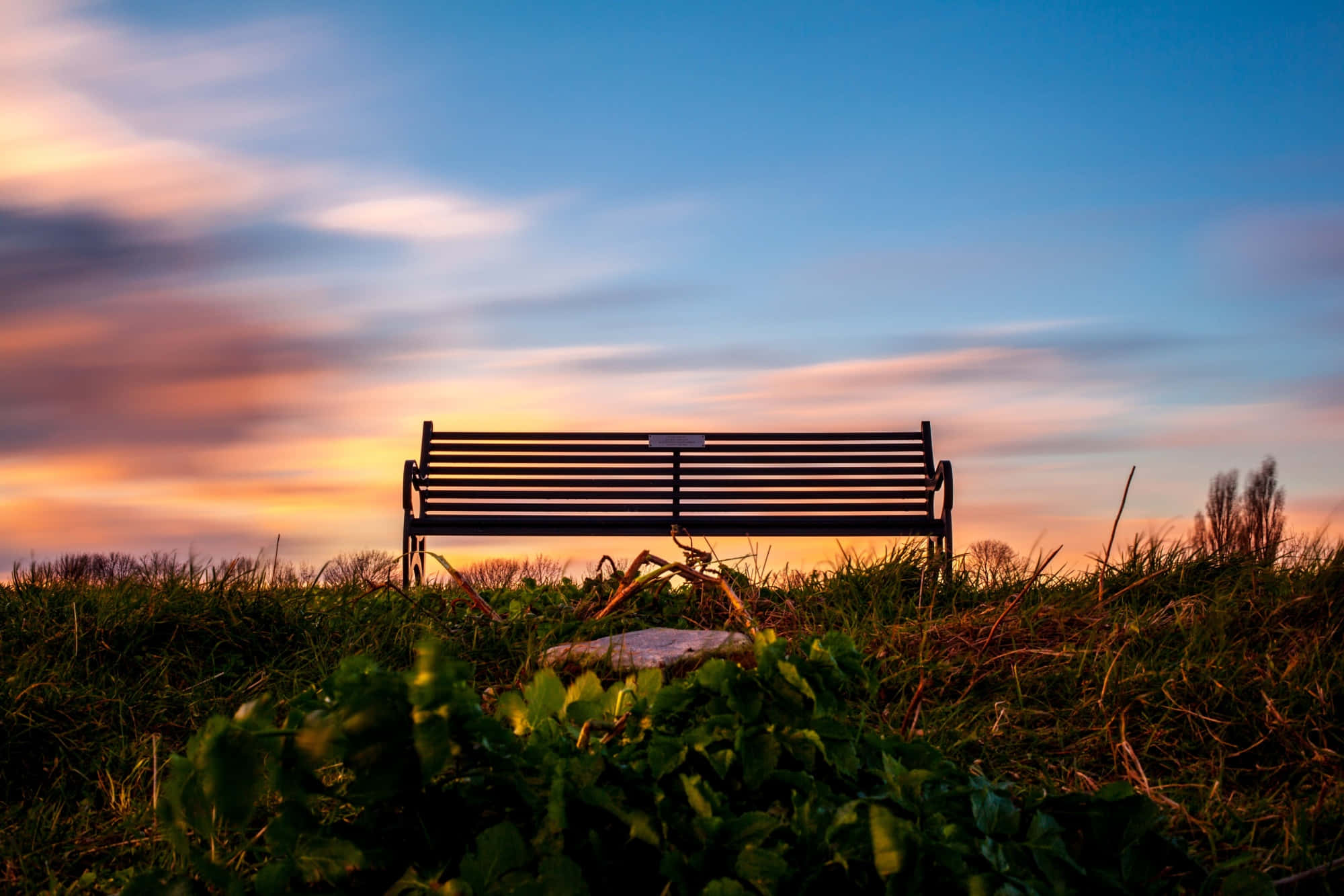 Bench 桌布 壁紙 — 一張孤獨的金屬長椅坐落在日落時分的草地上，在橙色和藍色雲彩條紋的鮮豔天空襯托下形成剪影。