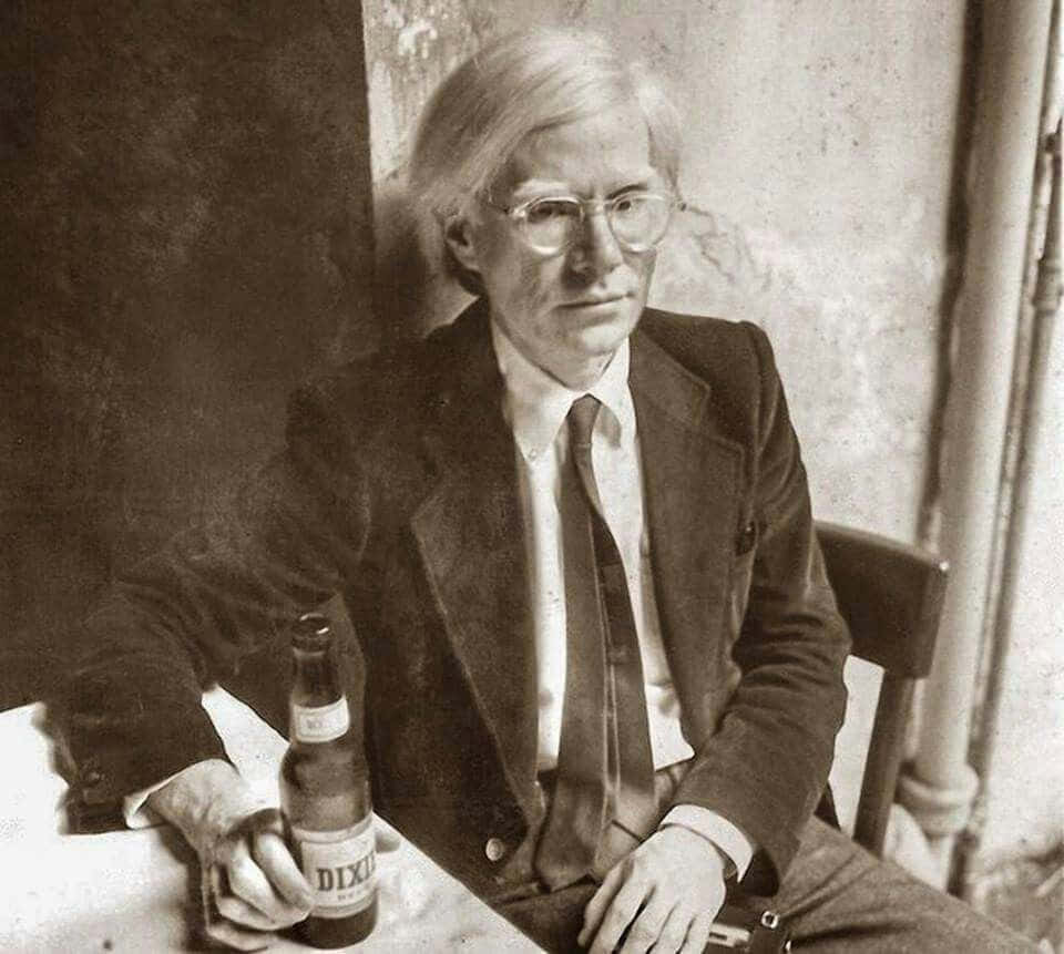 andy warhol 桌布 壁紙 — 一張安迪·沃荷的黑白肖像，他坐在桌子旁，手持啤酒瓶，表情嚴肅。