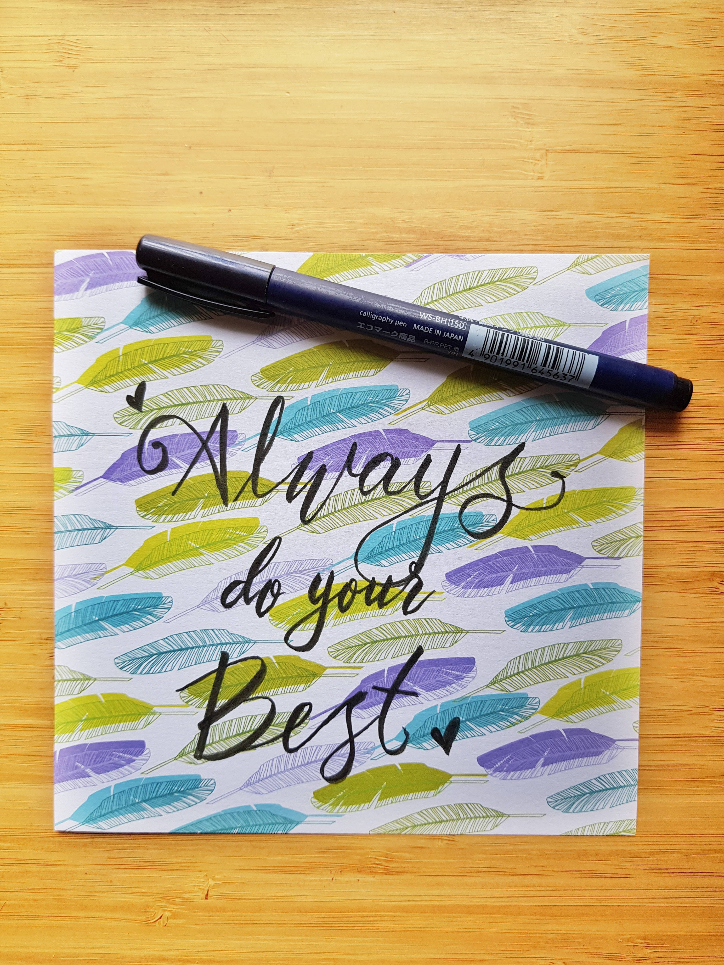 Best 桌布 壁紙 — 一張寫有「Always do your Best」優雅黑色字體的勵志便條，放在色彩繽紛的羽毛圖案紙上，旁邊有一支藍色記號筆，置於溫暖的木質表面上。