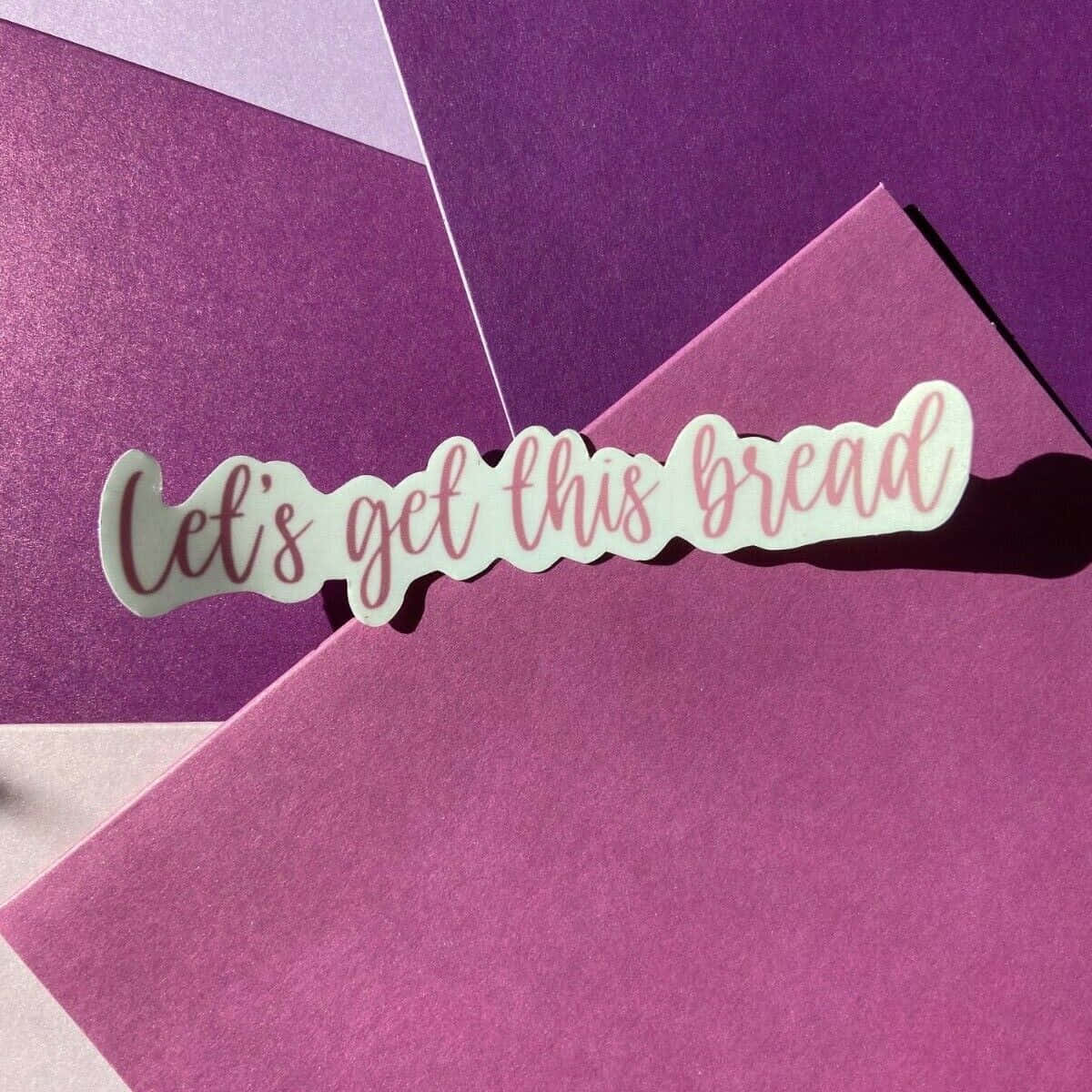  壁紙 — 一張寫有「Let's get this bread」的俏皮貼紙，放在柔和的日光下層疊的紫色紋理紙上。