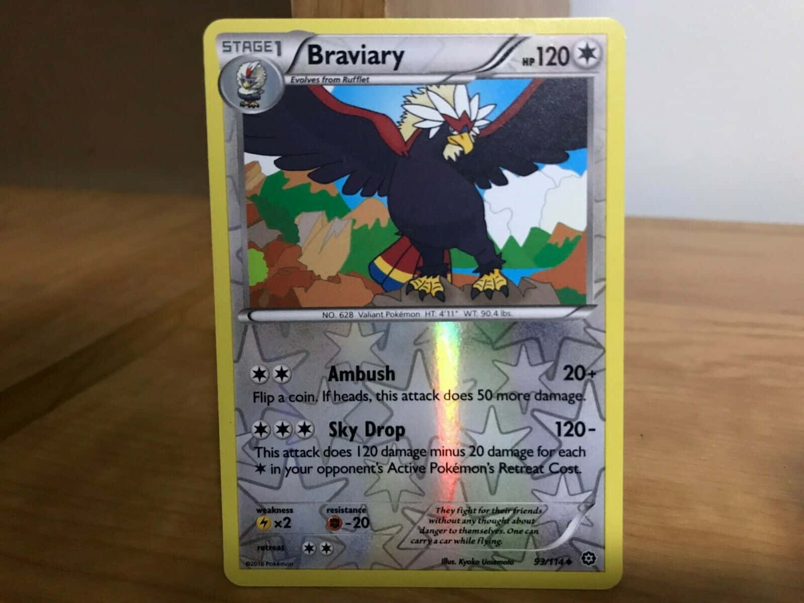 Braviary 桌布 壁紙 — 一張帶有全息光澤的勇士雄鷹寶可夢卡牌，在柔和室內燈光下放置於木質表面上。