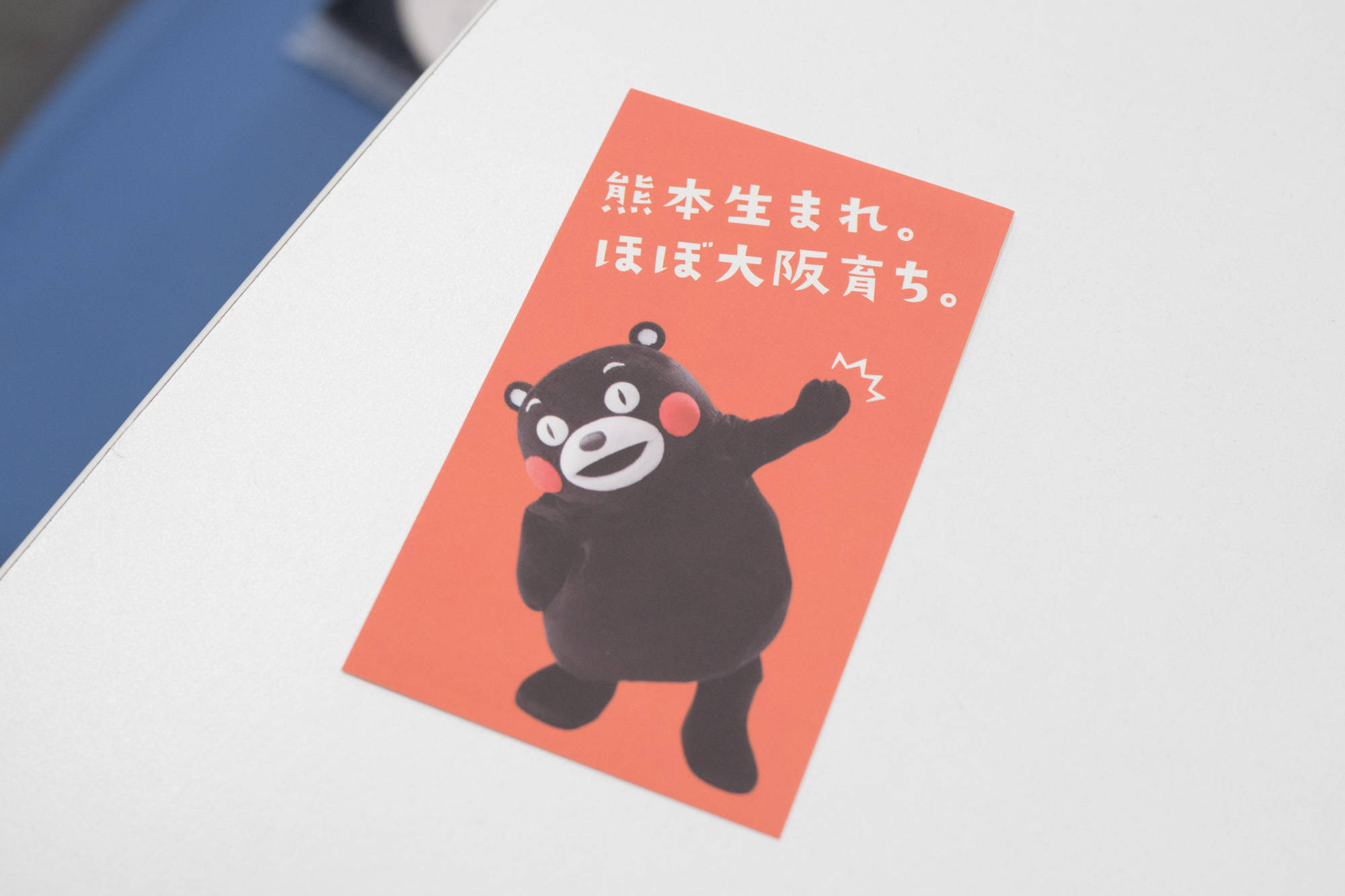 熊本熊 桌布 壁紙 — 一張帶有日文字樣的開心 Kumamon 貼紙，放在白色表面上，旁邊有一個藍色物體。