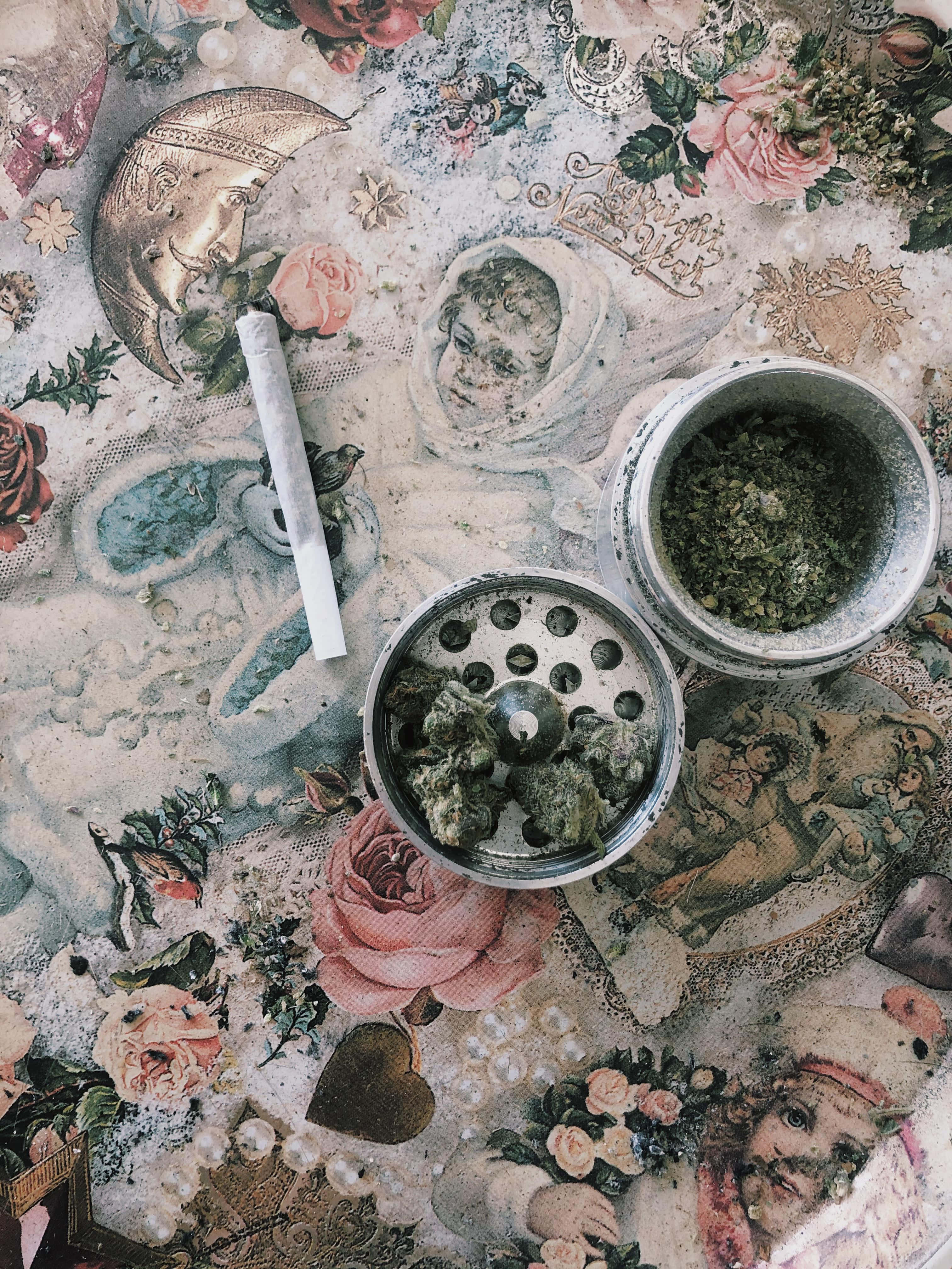Weed Blunt 桌布 壁紙 — 一張帶有玫瑰和小天使圖案的復古裝飾桌布，在夢幻而雜亂的美學中托著一個研磨器和一支大麻煙。