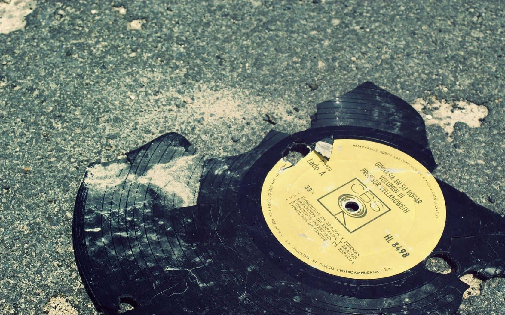 Vinyl Record 桌布 壁紙 — 一張帶有黃色標籤的破碎黑色黑膠唱片散落在粗糙的瀝青路面上。