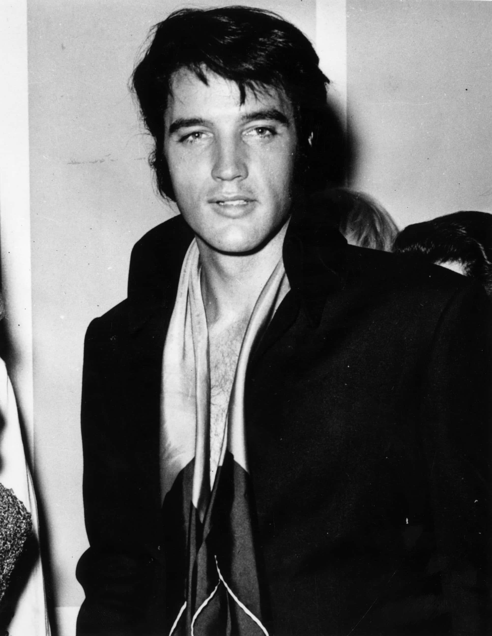 Elvis 桌布 壁紙 — 一張年輕貓王艾維斯·普里斯萊的黑白肖像，他看向鏡頭外，帶著淺淺微笑，身穿深色外套和圍巾。