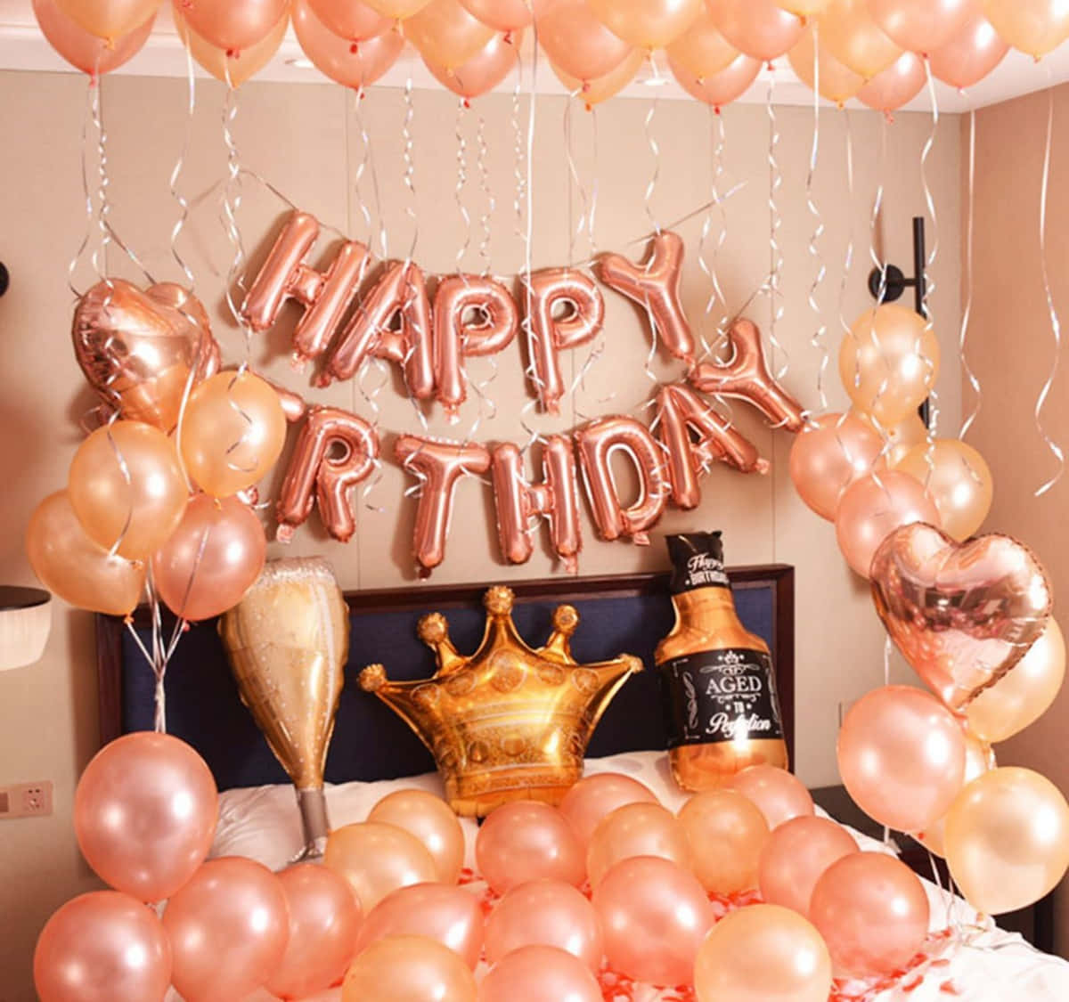 Birthday Celebration 桌布 壁紙 — 一張床，點綴著玫瑰金色氣球和「HAPPY BIRTHDAY」橫幅，周圍環繞著派對裝飾。