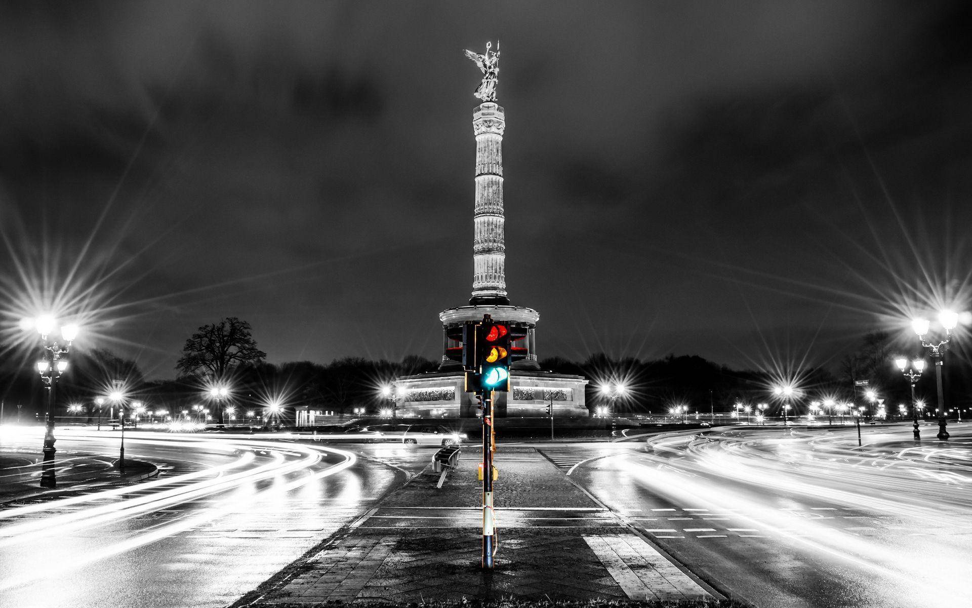 Berlin 桌布 壁紙 — 一張引人注目的黑白夜景照片，拍攝了一座高聳的紀念碑，頂部有一座發光的雕像，周圍環繞著汽車燈光軌跡和明亮的街燈。
