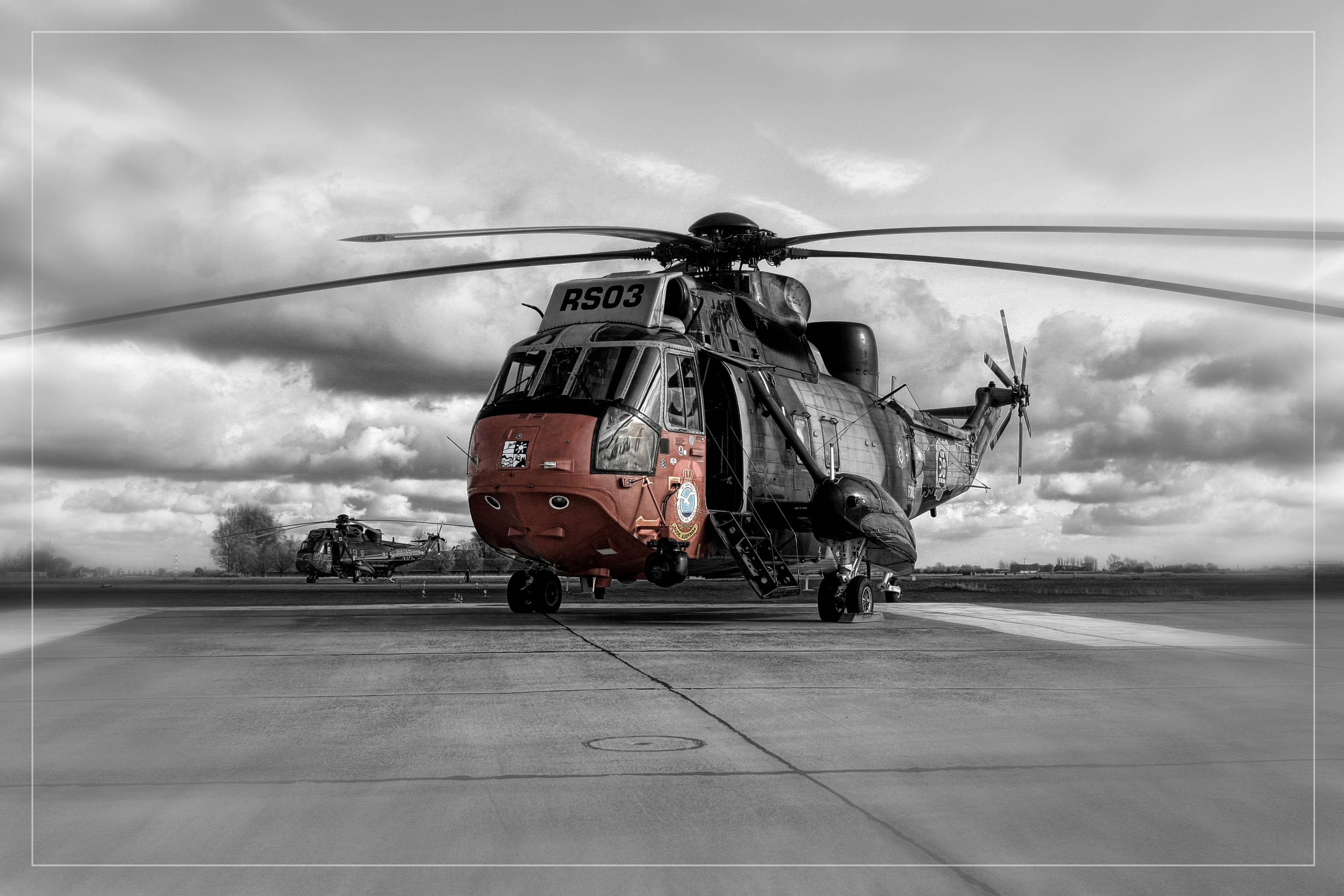 Helicopter 桌布 壁紙 — 一張引人注目的黑白照片，展示一架紅色軍用直升機停放在機場上，背景有另一架直升機，天空多雲。