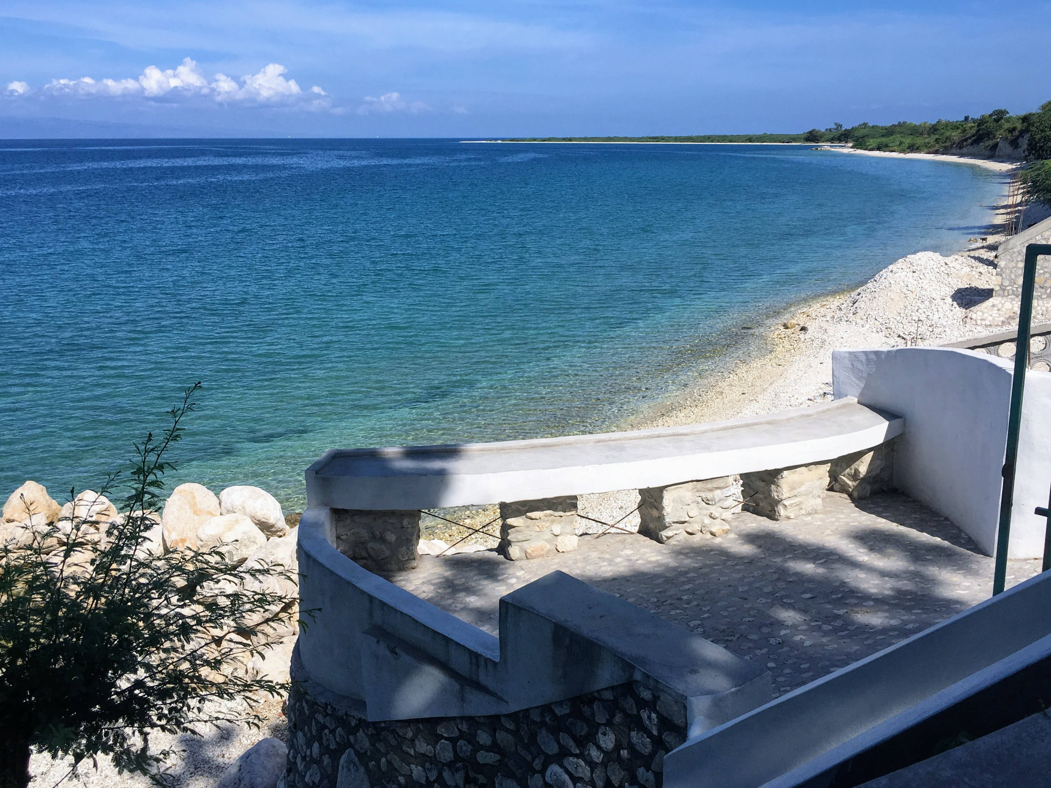 Haiti 桌布 壁紙 — 一張彎曲的混凝土長椅俯瞰著碧綠的海水與岩石海岸交匯，天空湛藍，白雲點點。