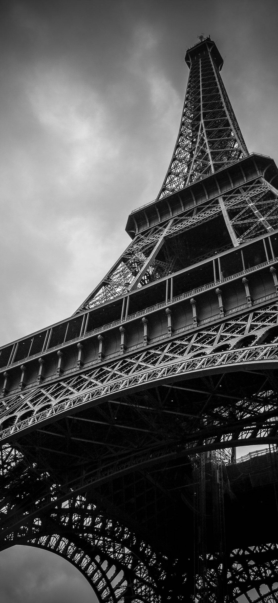 Eiffel Tower 桌布 壁紙 — 一張從低角度拍攝的戲劇性黑白照片，展現艾菲爾鐵塔複雜的鐵製格子結構，背景是陰雲密佈的天空。
