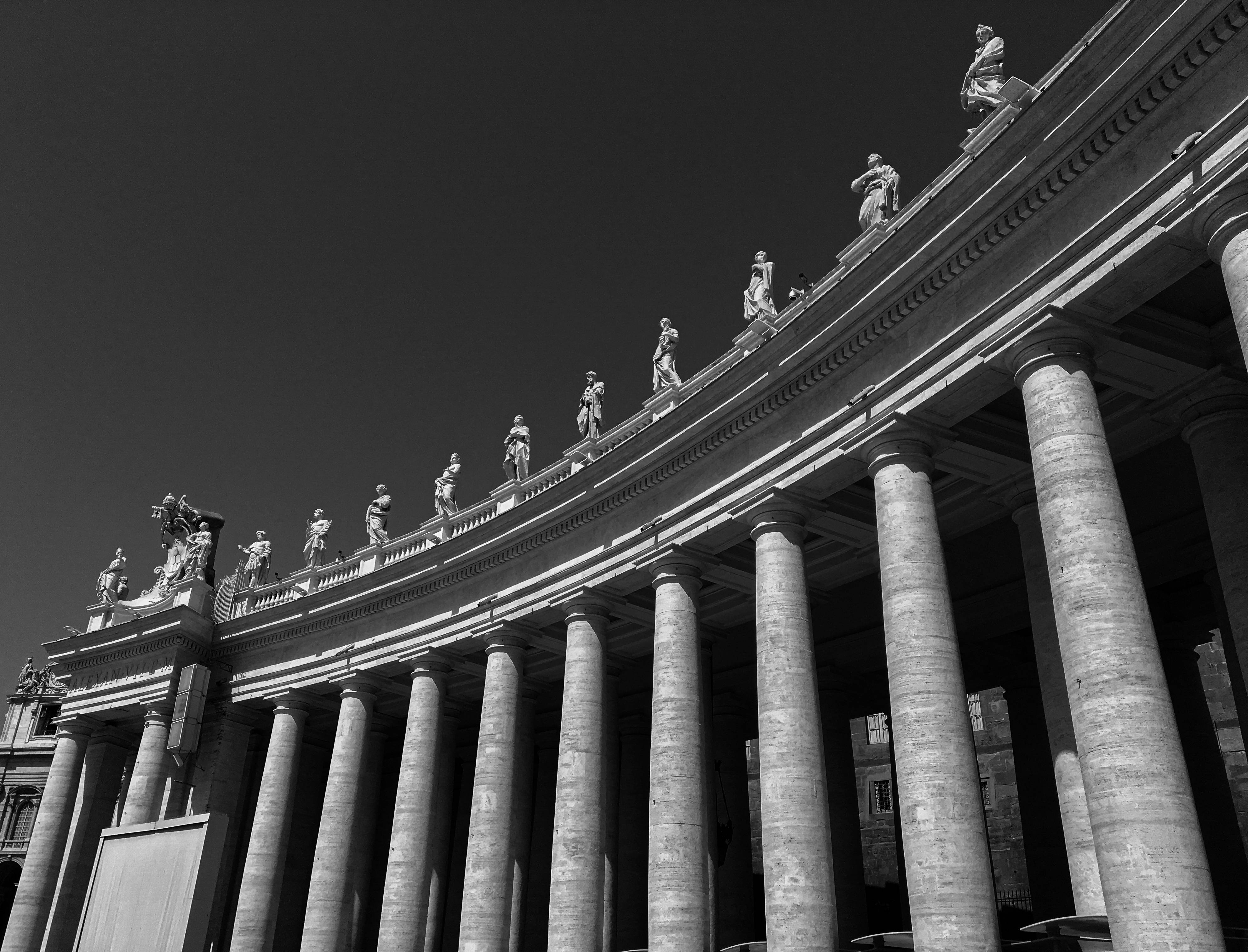 Vatican 桌布 壁紙 — 一張從低角度拍攝的戲劇性黑白照片，展示聖彼得大教堂的柱廊，柱頂橫楣上立有雕像，背景是 stark 的天空。