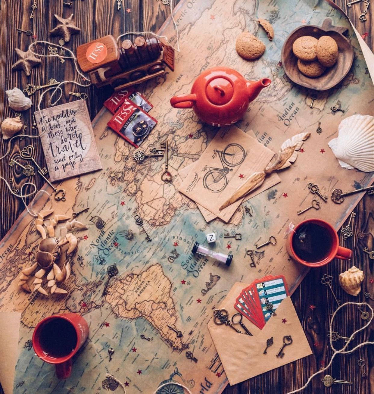 iPhone Travel 桌布 壁紙 — 一張復古世界地圖鋪在木桌上，裝飾著紅色茶壺杯、餅乾、貝殼和古董鑰匙，喚起旅行慾望。