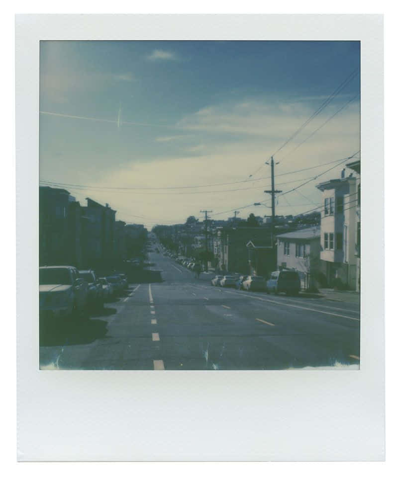 Aesthetic Polaroid 桌布 壁紙 — 一張復古拍立得風格的照片，展示了一條安靜的城市街道，兩旁停著汽車和住宅建築，天空多雲。